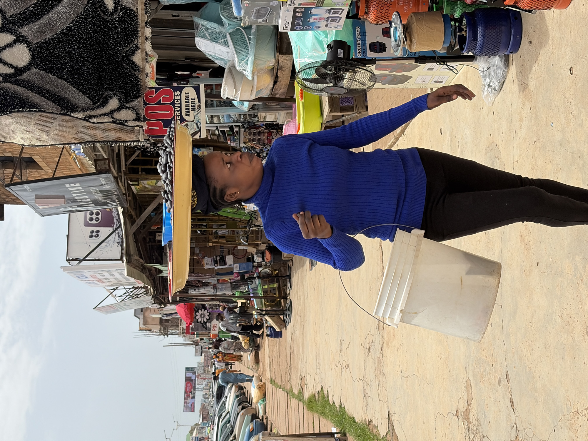 A young woman wearing a blue, long-sleeved shirt and black pants stands in an outdoor market in Jos, Nigeria. She is holding a white plastic bucket. Behind her, stalls are set up with various goods, including fans, gas cylinders, and packaged items. Signs are visible, with text that reads "POS SERVICES AVAILABLE HERE" and "ORCHID TOYS COLOR:". The market appears to be busy, with other people and parked vehicles visible in the background. The ground is paved with concrete. The lighting suggests it is daytime.