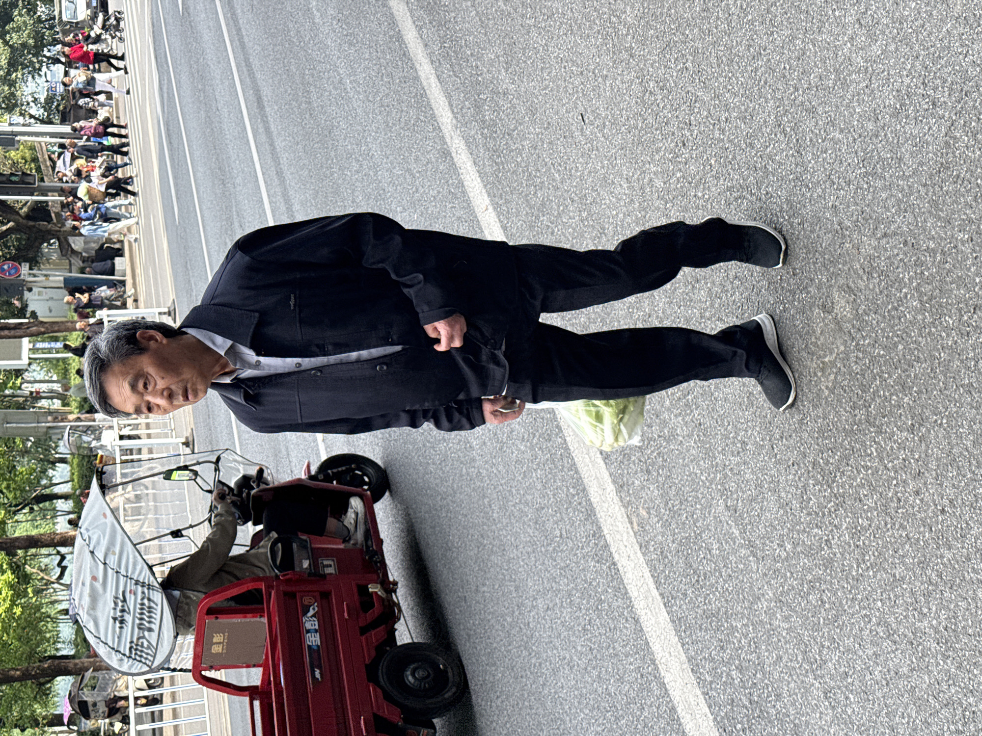 An Asian man wearing a dark suit and dark sneakers stands in the right-hand lane of a gray asphalt road with white lane markings. The man is facing the camera with a bag of vegetables at his side. Adjacent to him, on the left side of the frame, is a red motorized tricycle with a white awning. An individual is seated in the tricycle. In the background, across the road, is a crowd of people walking on a sidewalk. Behind them are trees and buildings. The image was taken in Jiangjin, China.