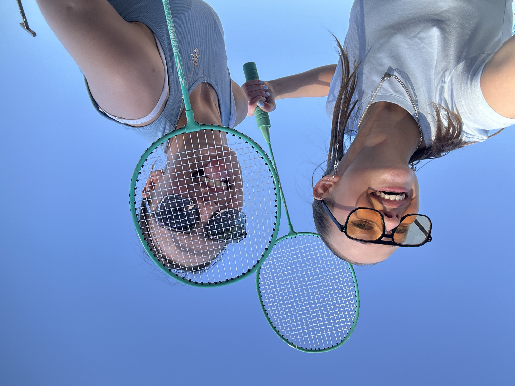 Two young women are playing badminton in Khimki, Russia. The image is taken from a low angle, looking up at them against a bright blue sky, suggesting it is daytime with clear weather. Both individuals are upside down in the frame.
The woman on the left is wearing a light blue tank top and appears to have sunglasses on. She is holding a green badminton racket. The woman on the right is wearing a light blue t-shirt and oversized sunglasses with orange lenses. She is also holding a green badminton racket. Both women are smiling, indicating a fun and playful mood.
The background is a clear, uninterrupted blue sky, offering no specific location cues other than the general assumption of an outdoor setting suitable for badminton. The image captures a moment of leisure and enjoyment between the two friends. There is visible text on the racket of the woman on the left, which appears to say "HONGZHAN 20" and possibly more that is cut off.