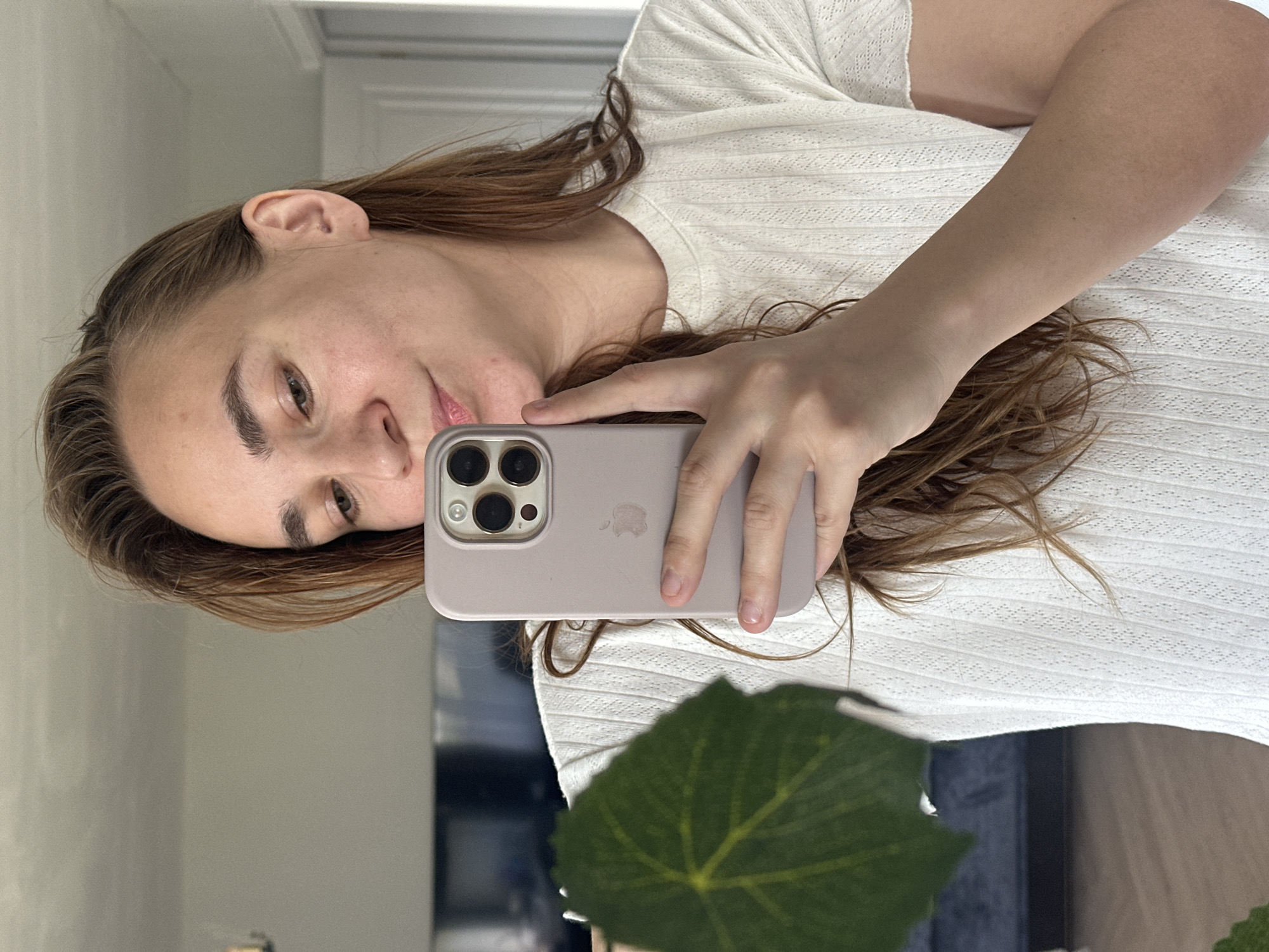 A fair-skinned person with light brown hair is holding a smartphone, likely an iPhone, in front of their face to take a selfie. The phone is in a lavender or light purple case. The person is wearing a white shirt with a textured pattern. A large green leaf is partially obscuring the lower portion of the view. The background appears to be a reflection in a mirror, showing a wall and a dark object or piece of furniture. The photo does not show any location in Kharnavy, Russia.