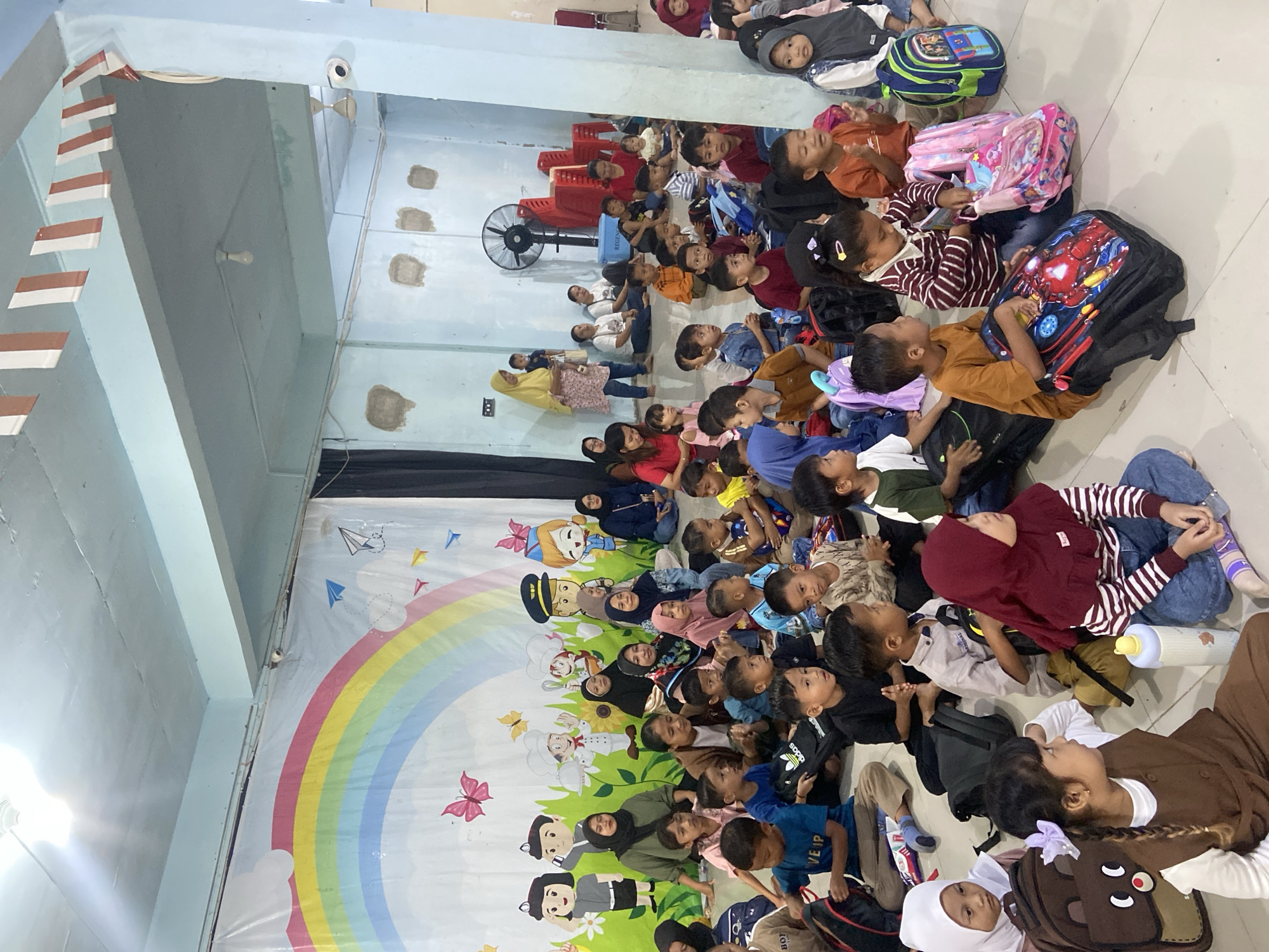 The image depicts an indoor gathering of approximately 50 to 60 children and adults, likely in a community hall or classroom in Tanjung Gading, Indonesia. The scene is brightly lit, with a large, colorful mural of a rainbow, clouds, and cartoon characters decorating the back wall. The floor is a light-colored tiled surface.
In the foreground and midground, numerous children are seated on the floor, facing towards the back wall and an unseen focal point. They are of various ages, with many appearing to be preschoolers or early elementary school students. Most are dressed casually, and many have backpacks or school bags placed around them. Some children are engaged with their bags, while others are looking forward or interacting with each other.
Adults are also present, interspersed among the children and seated on the floor or on a raised platform at the back of the room. Several women are wearing hijabs, common in Indonesia. The overall atmosphere appears to be one of a children's event, possibly an educational program or a celebration.
The environment includes a ceiling with exposed beams and lighting fixtures. There is a large fan mounted on the wall in the background, and several stacked red chairs are visible near the back. Security cameras are mounted on the walls. Decorative red and white striped flags are strung across the ceiling. The walls are painted a light blue, and there are some visible patches of plaster on the walls, suggesting ongoing maintenance or a rustic aesthetic. The overall scene suggests a humble but functional community space.