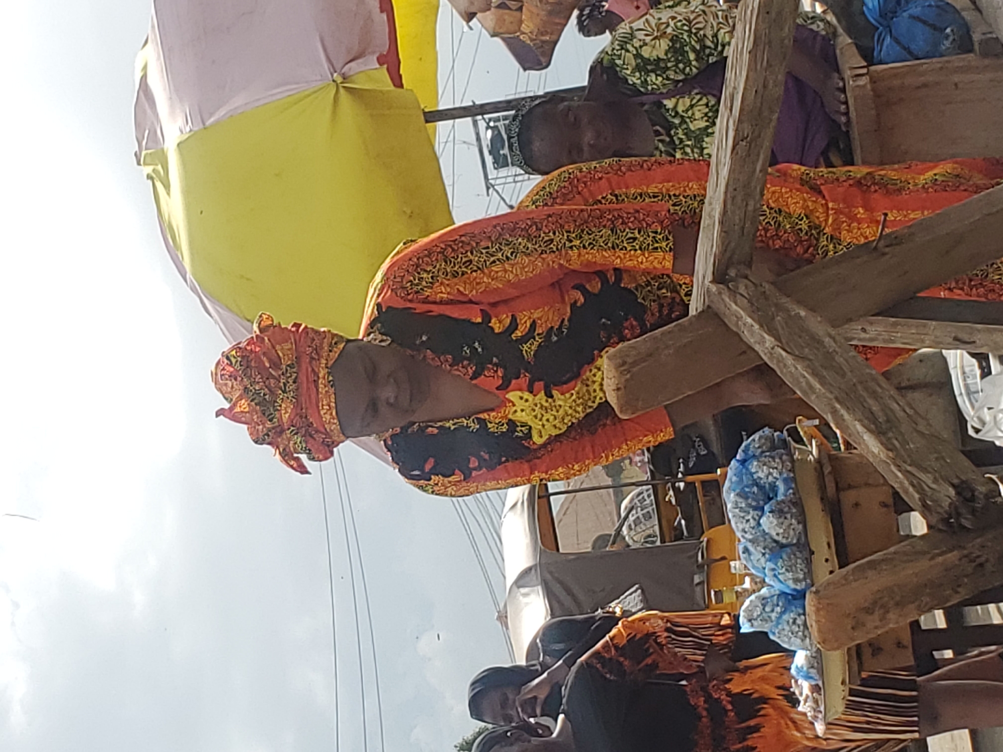 A woman wearing a colorful headwrap and a vibrant orange and black patterned dress is depicted at what appears to be a market in Gwong, Nigeria. She is bent over, looking down at a tray of items. The tray, supported by a wooden structure, holds several blue-wrapped parcels containing what looks like nuts or small food items. Behind her, another woman in a green and white floral dress is partially visible, sitting down. In the background, a yellow and white umbrella provides shade, and a vehicle, possibly a tricycle or auto-rickshaw, is parked nearby. The scene suggests a bustling outdoor marketplace with vendors and shoppers. The sky is overcast, indicating a cloudy day. The overall impression is of everyday life in a Nigerian town.