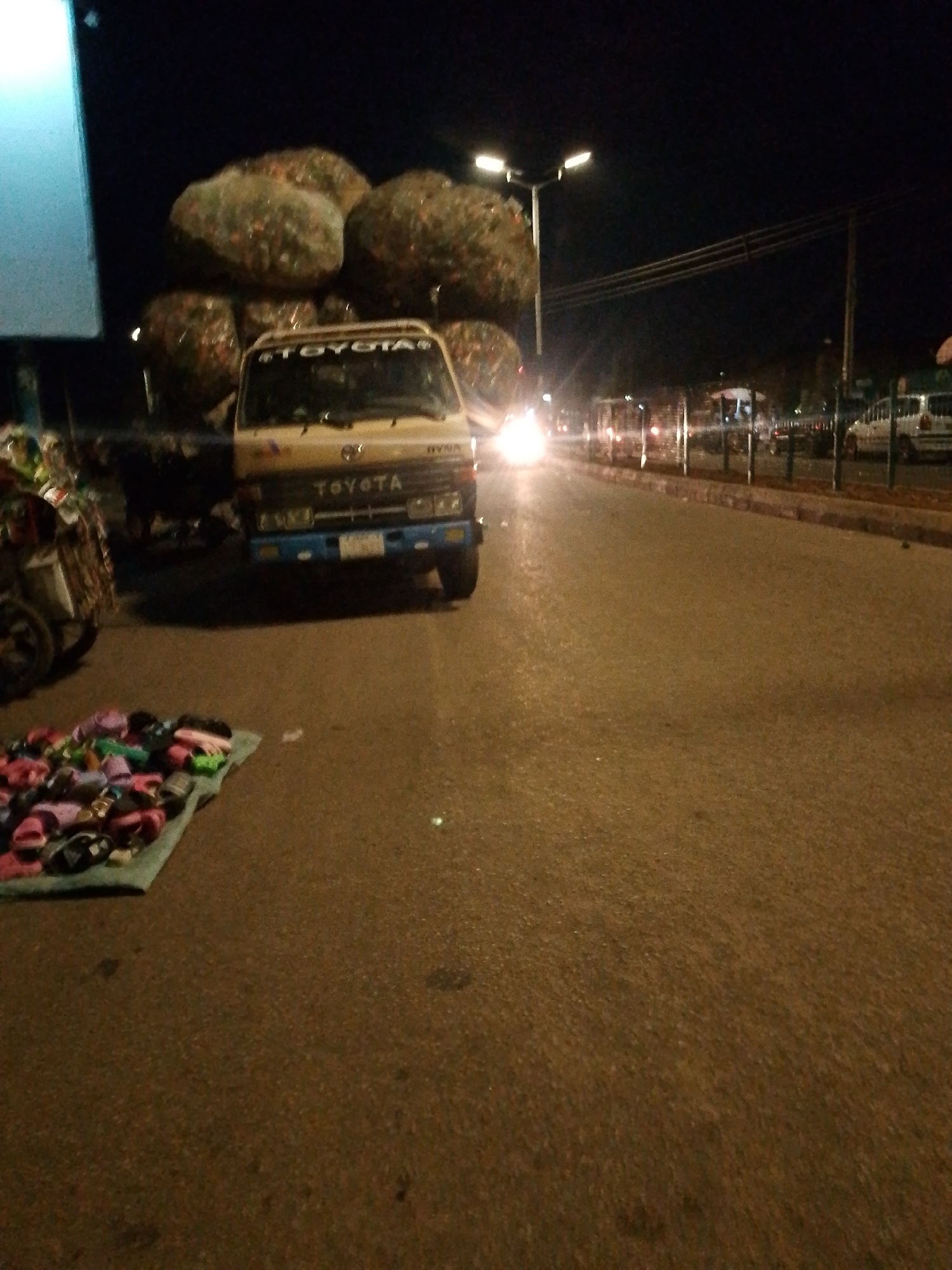 The image depicts a nighttime scene along a road in Bukuru, Nigeria. A beige Toyota truck is prominently parked on the left side of the road, heavily loaded with large, irregular bundles that appear to be sacks of goods or possibly waste. The brand name "TOYOTA" is clearly visible on the truck's grille and also as a sticker above the windshield. The model name "Dyna" is also present on the front of the truck.
In the foreground, to the left of the truck, a diverse collection of colorful sandals and slippers is neatly laid out on a light-colored mat directly on the asphalt, indicating a roadside stall or vendor. A handcart or trolley is partially visible further left.
The scene is lit by various artificial light sources. Bright headlights from an oncoming vehicle are visible further down the road on the right, creating a strong glare. Streetlights with twin fixtures illuminate the road in the distance. The road itself is paved and appears dark, wet, or well-used. Metal barriers or fences line the right side of the road, separating it from what appears to be a parallel path or another road. The overall impression is of a bustling but dimly lit urban or market environment during the night, with commercial activity underway. No individuals are visible in the frame.