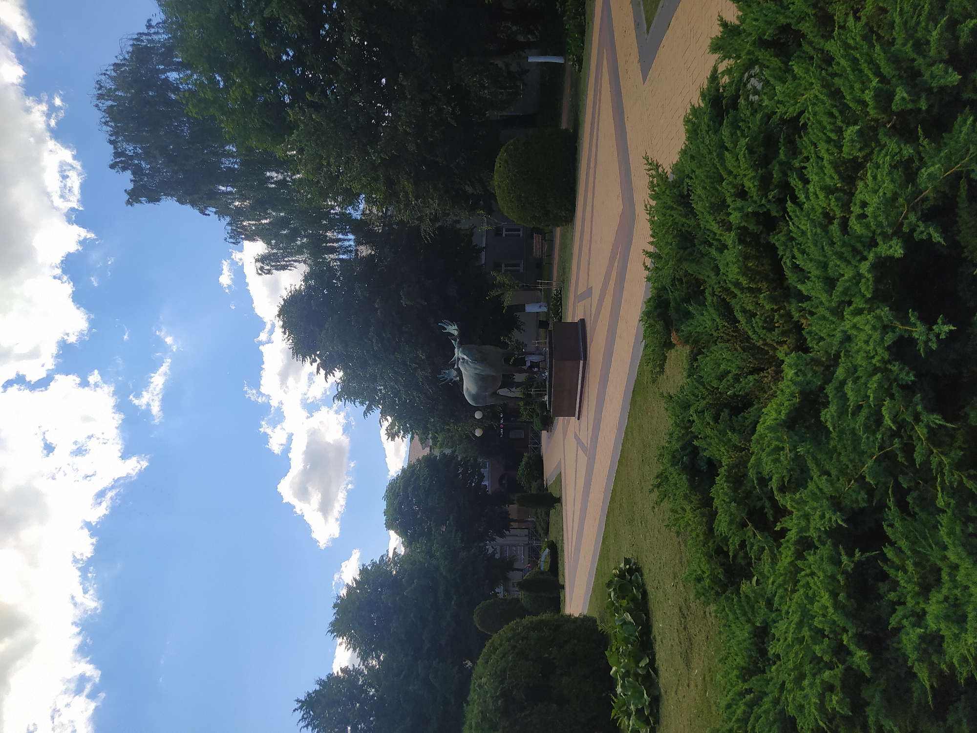 The image captures a serene outdoor scene with a clear blue sky dotted with fluffy white clouds. Lush green trees and manicured bushes dominate the foreground and middle ground. A paved pathway, possibly in a park or public square, is visible, with geometric patterns of light and dark yellow or beige tiles.
In the middle distance, a bronze sculpture of a bull stands on a pedestal. The sculpture is detailed and appears to be a significant landmark. Behind the sculpture, buildings with multiple windows are visible, suggesting an urban or semi-urban setting.
The overall impression is one of a pleasant day, likely in the late morning or early afternoon given the bright sunlight casting shadows. The weather appears to be fair and warm, ideal for outdoor activities. The presence of trees, well-kept greenery, and a public walkway suggests a well-maintained public space.
The location is identified as Gusev, Russia. While no specific activity is depicted, the scene conveys a sense of peace and tranquility. The focus is on the natural elements, the architectural details of the pathway, and the central sculpture. No visible text is present in the image.