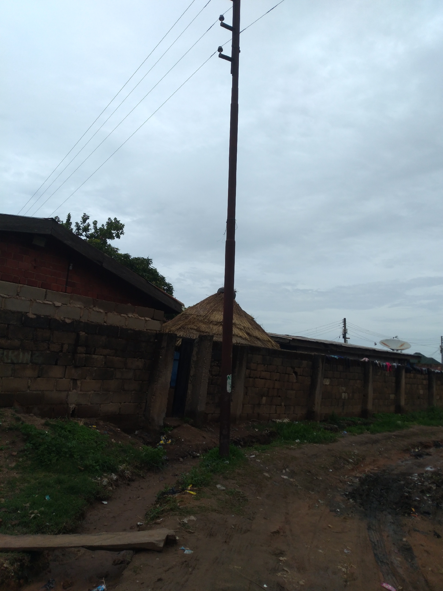 A utility pole stands vertically in the center foreground of the image, its dark brown surface marred by a vertical crack. Overhead, four power lines stretch diagonally across the cloudy, pale gray sky. To the left of the pole, a section of a reddish-brown brick building with a dark, sloped roof is visible, partially obscured by a low, rough-hewn stone wall. This wall, constructed from irregular, light-colored bricks, runs horizontally across the middle ground, extending to the right. Behind the wall, a thatched-roof structure with a conical shape is partially visible, suggesting a traditional dwelling. Further to the right, the stone wall continues, and a white satellite dish with a circular receiver is mounted on a pole. The ground in the foreground is uneven and muddy, with patches of green grass growing to the left and a scattering of debris, including plastic bags and what appears to be a wooden plank spanning a ditch. The overall impression is of a somewhat disheveled urban environment in Jos, Nigeria, with a mix of modern and traditional structures under an overcast sky.