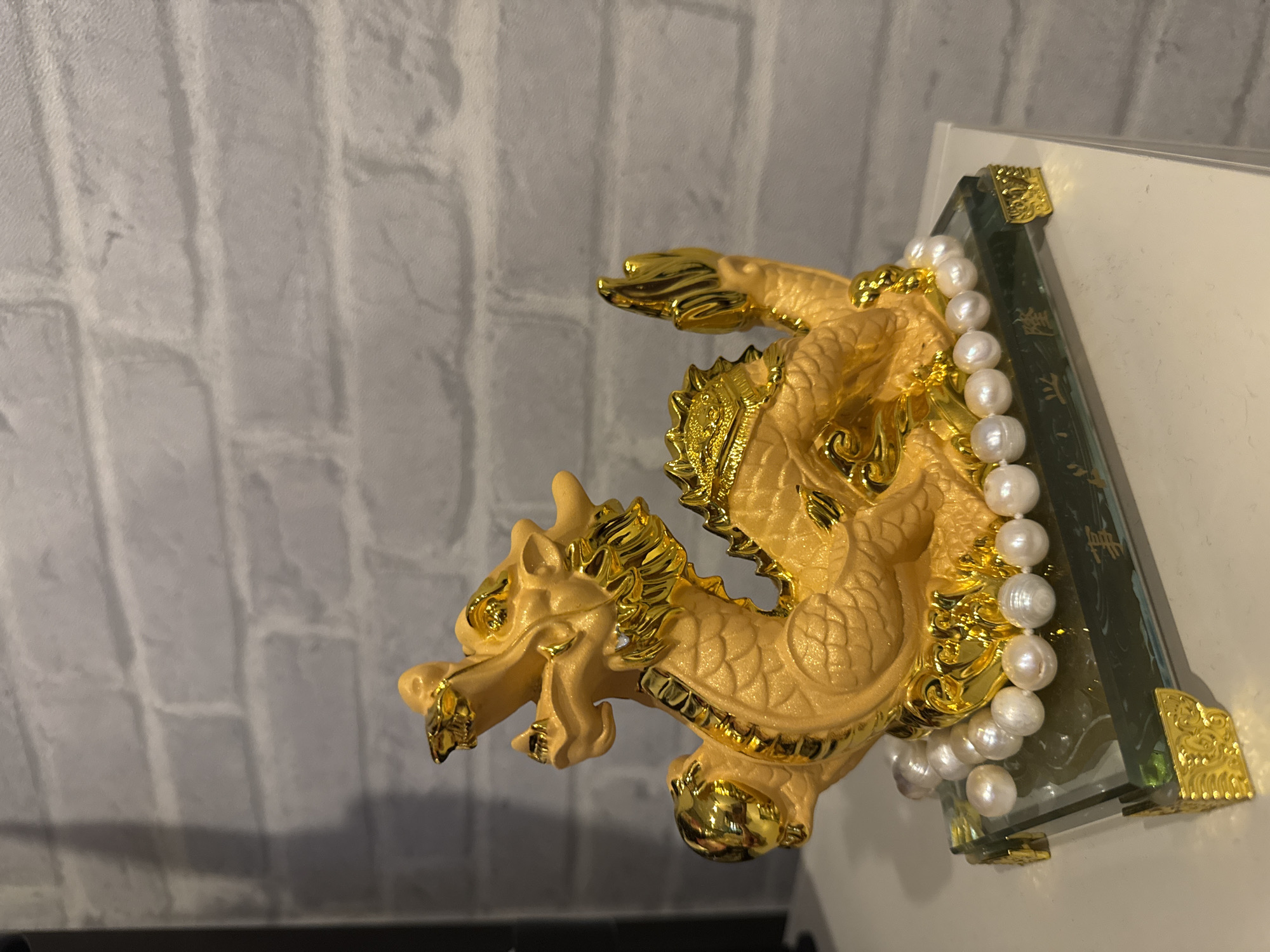 The image displays a golden-colored, ornate dragon figurine as its central subject. The dragon, depicted in an dynamic pose, features intricate details including textured scales, a flowing mane, and pointed claws, rendered in both a glossy gold and a slightly lighter, matte-gold finish. Its mouth is open, giving an impression of roaring or breathing.
Draped around the dragon's body and resting against a supporting structure is a strand of white, irregularly shaped (baroque) pearls. The pearls exhibit a soft sheen. The dragon and pearls are positioned against a clear glass or acrylic block that has dark green or black sections. These sections are adorned with faint golden text, likely Asian script, arranged in vertical lines, though the characters are not legible. The corners of this glass block are further embellished with small, ornate golden metallic brackets.
The background is a light grey or off-white wall featuring a textured brick pattern, indicating an indoor setting. The lighting creates highlights on the metallic and pearlescent surfaces. The object appears to be a decorative item, possibly symbolizing prosperity or good fortune, consistent with cultural motifs often found in locations like Bangkok, Thailand. There are no people visible in the image, and no specific activity is taking place beyond the display of the figurine.