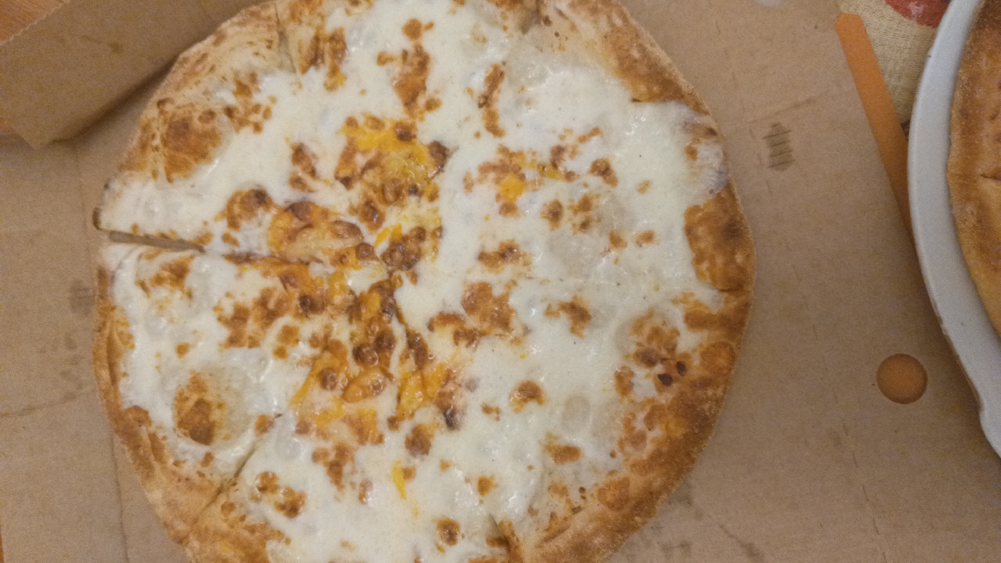 The image shows a close-up, overhead view of a partially eaten pizza, likely a white pizza due to the absence of tomato sauce. The pizza is on a brown cardboard pizza box, which is placed on a textured surface, possibly a table. The pizza is round and has a golden-brown crust with some charring. It is topped with melted white cheese, which has some golden-brown baked spots, and appears to have some type of shredded cheese or spice mixed in. The pizza has been cut into several slices, with at least two slices already removed. To the right of the main pizza, a portion of another pizza in a white dish is visible, also appearing to be a white pizza. The overall lighting is dim and appears to be from an overhead source, creating some shadows. There are no people or animals visible in the image. The location within Mytishchi, Russia, cannot be determined from the visual information provided in the image.