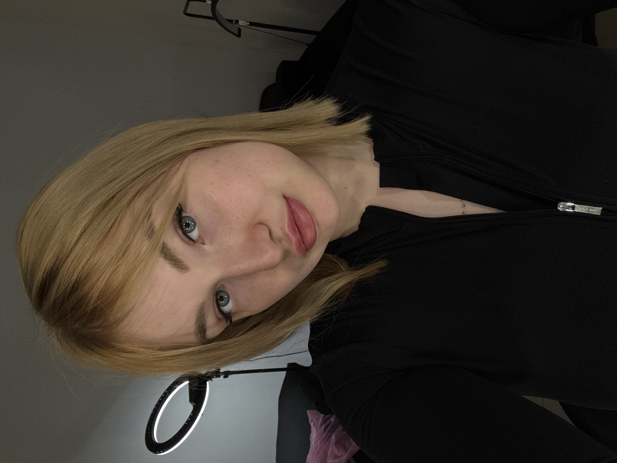 A young woman with blonde shoulder-length hair and blue eyes is shown in a close-up, head-and-shoulders shot. She is wearing a black zippered top and has light makeup on. In the background, a ring light is visible on the left side, and a standing lamp is on the right. The environment appears to be indoors, possibly a room with dark walls. There is a hint of a pink object on a dark surface to the left of the woman. No specific details indicate the location in Moscow, Russia.