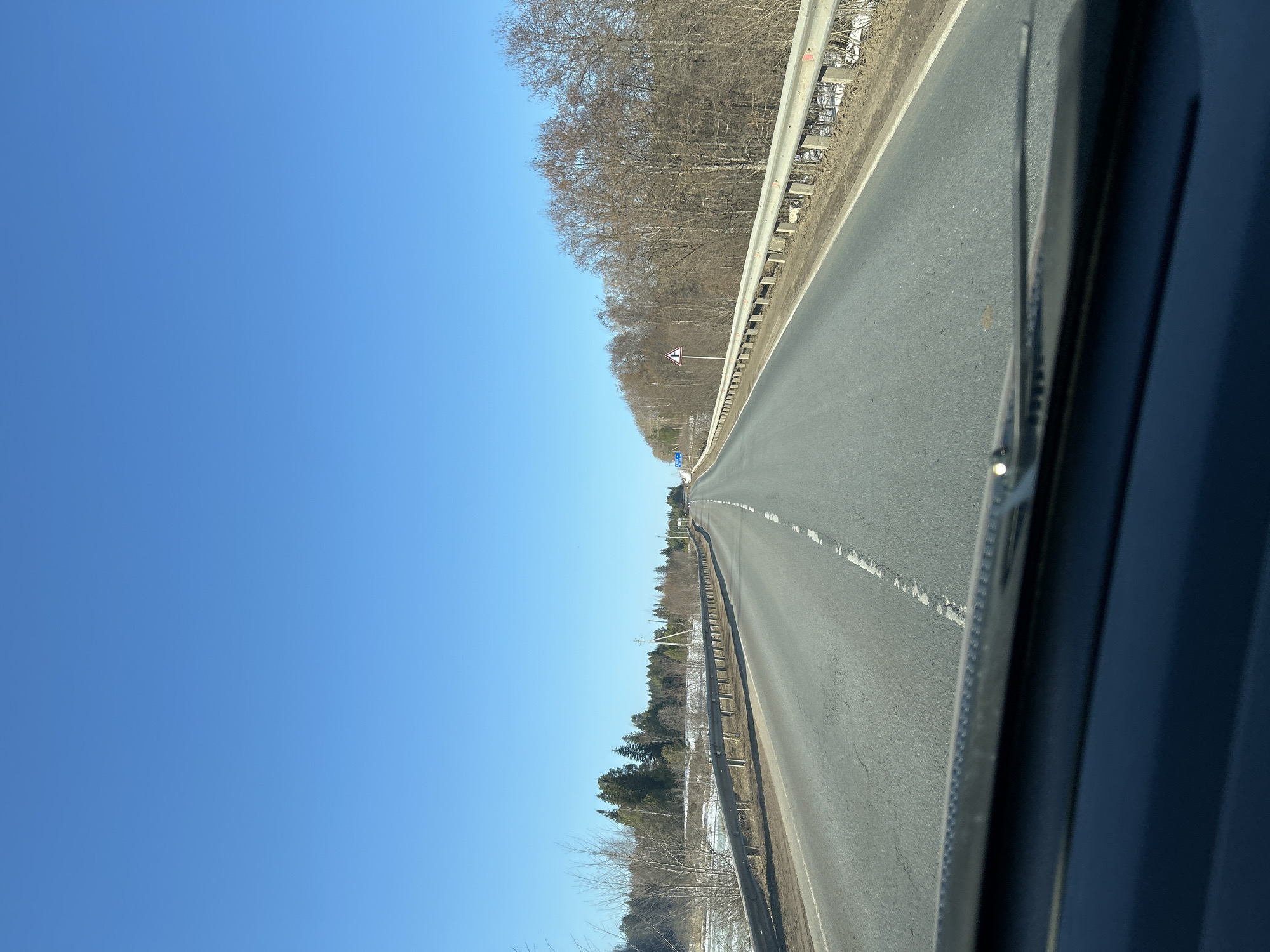 The image shows a straight, paved road in the vicinity of Kamennyi Kliuch, Russia. The road has lane markings and guardrails on either side. Trees line the roadside. The sky is clear and blue. The image is taken from inside a vehicle, with part of the vehicle visible in the foreground. The absence of other vehicles or people suggests a sparsely populated area or time of day with low traffic. Bare trees indicate either early spring or late autumn. There is evidence of snow, suggesting that it is either late winter or early spring.