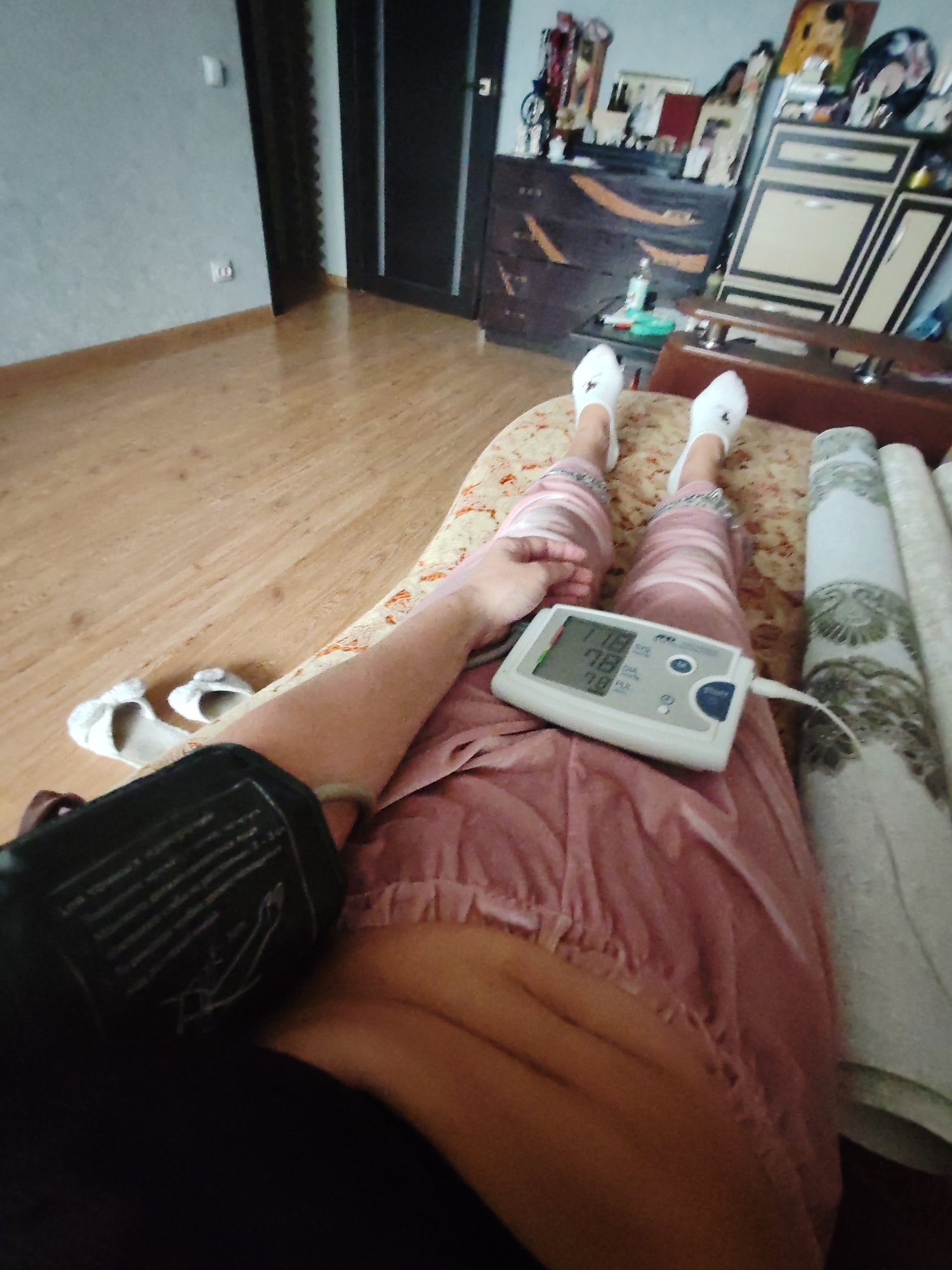 A person lies on a bed in a room, taking their blood pressure. The person is wearing white socks and pink satin-like pants. A blood pressure monitor is on their lap displaying a reading of 118 over 78, with a pulse of 78. The blood pressure cuff is on the person's upper arm. In the background, there is furniture, a door, and a wooden floor. There is no evidence suggesting that this photo was taken in Petropavlovsk-Kamchatsky, Russia.