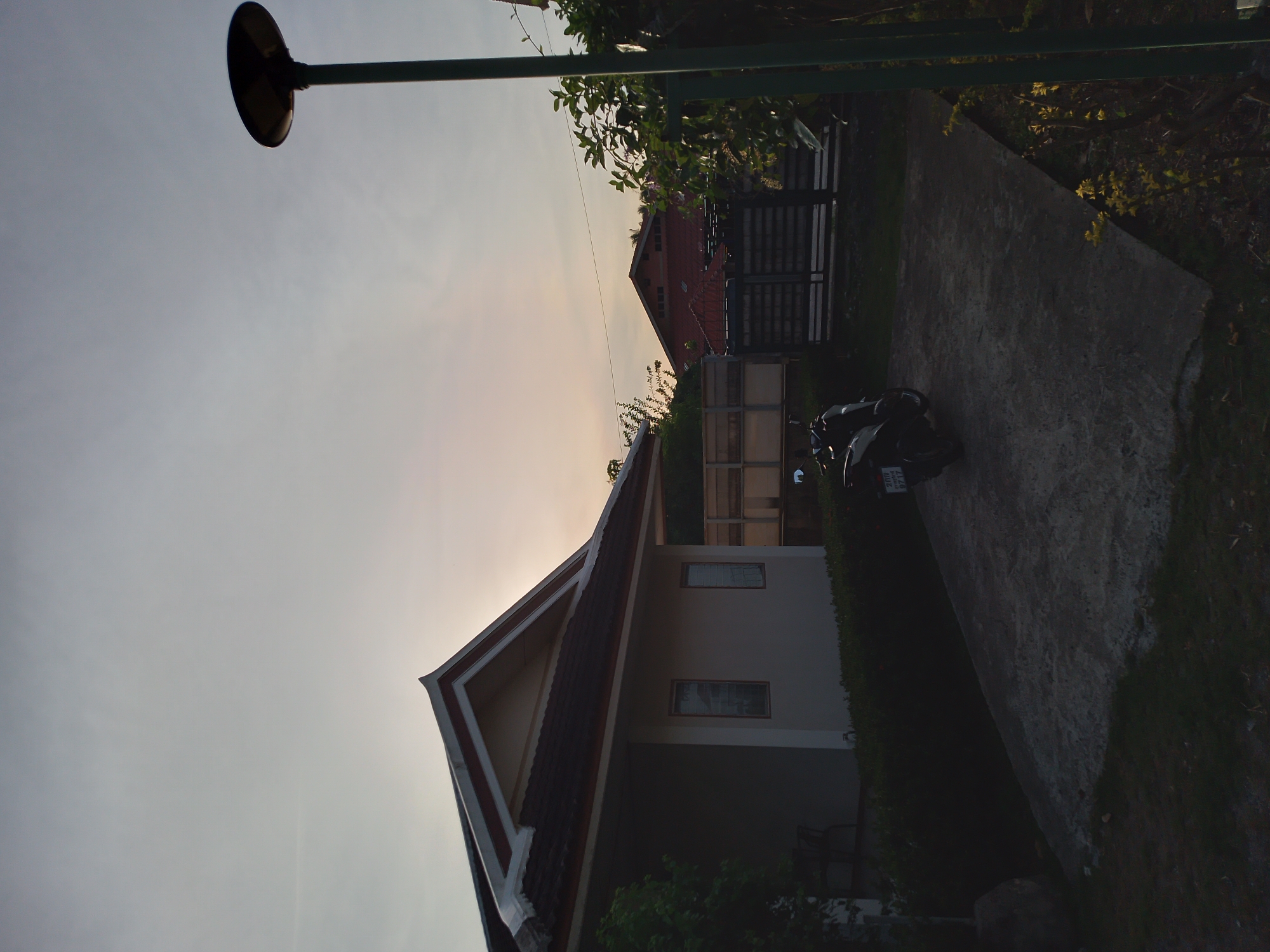 This image depicts an outdoor scene at dusk in Baan Mae Nam, Thailand. In the foreground, a dark-colored scooter is parked on a concrete driveway. To the left of the scooter is a single-story building with a light-colored exterior and a dark-tiled roof that features a distinctive upward-curving gable. Two windows are visible on the side of the building. A tall, dark-colored lamp post with a circular fixture stands to the right of the scene, partially obscuring some foliage and another building in the background. The background also shows a red-roofed structure with a fence and more vegetation. The sky is a pale, overcast gray, suggesting either early morning or late evening. There are no people visible in the image.