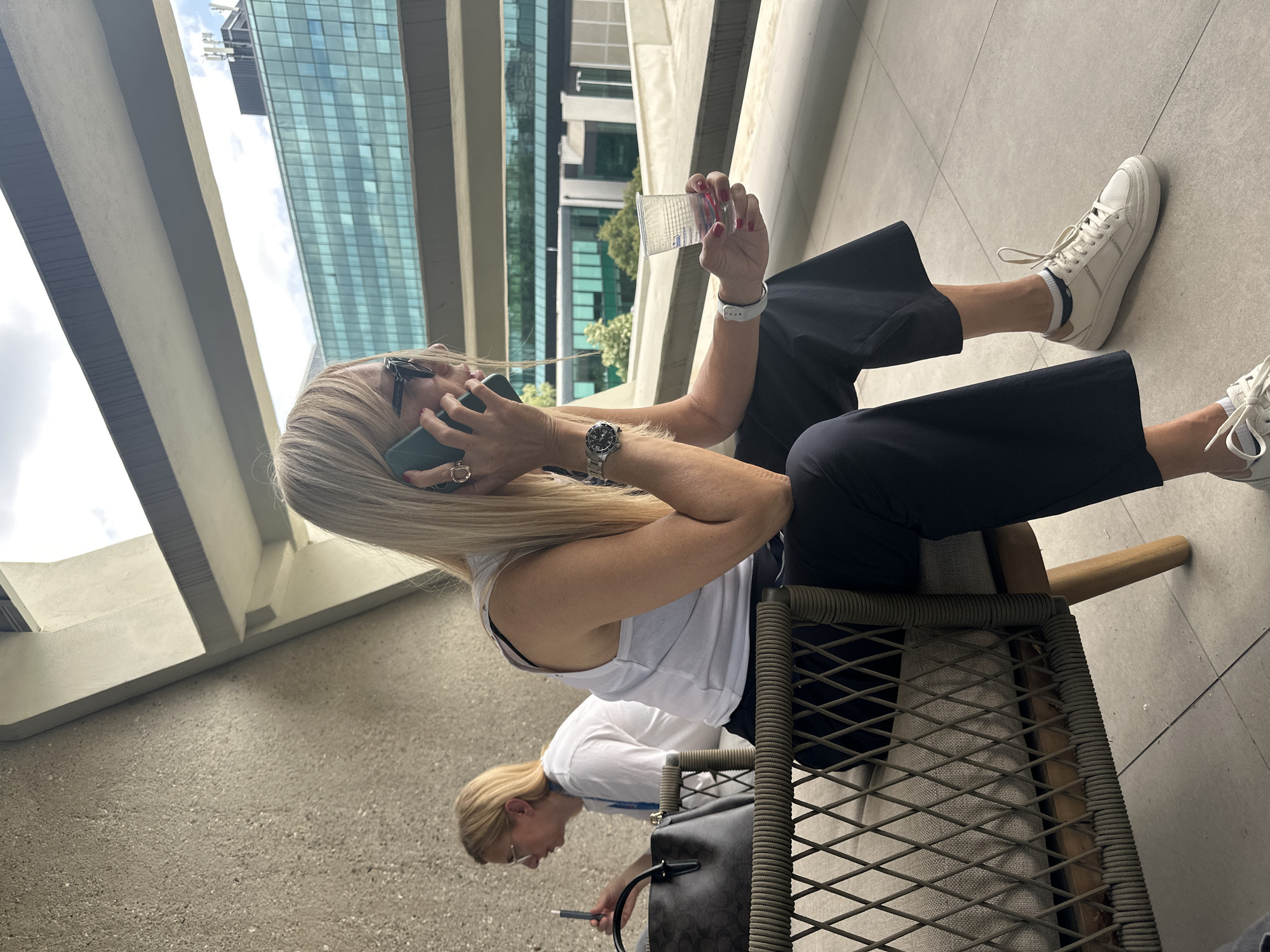 The image shows two women seated on a balcony, likely in Belgrade, Serbia given the architectural style. One woman, dressed in a white tank top and dark culottes, is on a mobile phone. She holds a clear plastic cup in her other hand. The second woman, dressed in a white shirt, is sitting and looking at her phone. Both women are wearing sneakers. Behind them is a modern building with large glass windows. The balcony has a grey concrete floor. The woman on the phone is seated on a chair with a woven rope seat.