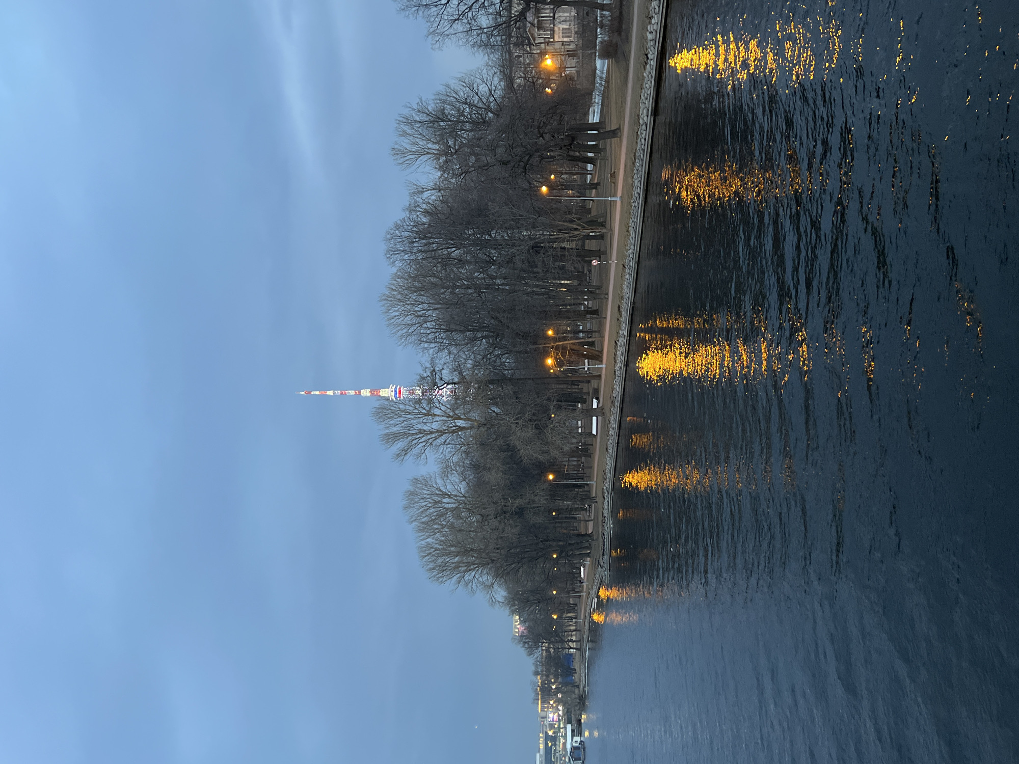 The image depicts an evening or dusk scene along the Malaya Nevka river in Saint Petersburg, Russia. The prominent Saint Petersburg TV Tower, illuminated with red, white, and blue segments, rises above the tree line in the background, situated on Aptekarsky Island.
In the foreground, the dark, rippled surface of the river occupies the lower half of the frame. It reflects the warm orange light from multiple streetlights positioned along the opposite bank, creating distinct vertical streaks of light on the water.
The right bank, likely part of Aptekarsky Island, features a structured embankment composed of stones or concrete. Immediately above this, a bare dirt or gravel path runs parallel to the river. A park area is visible, populated by numerous tall, bare deciduous trees, indicating a late autumn or winter season. Several streetlights with warm orange illumination line the path, and a few park benches are discernible beneath the trees. Small patches of white snow are present on the ground. A small white signpost with a red circular top is located near the path on the right.
In the mid-background, through and behind the trees, various urban structures are faintly visible, including a multi-story building with dark windows on the far right. On the distant left bank, across the river, additional city lights and faint building outlines can be observed.
The sky is overcast and exhibits a consistent blue-grey hue. A faint crescent moon is visible in the upper left quadrant of the sky. No individuals are present in the frame, and no discernible actions or interactions are occurring. The scene is static.