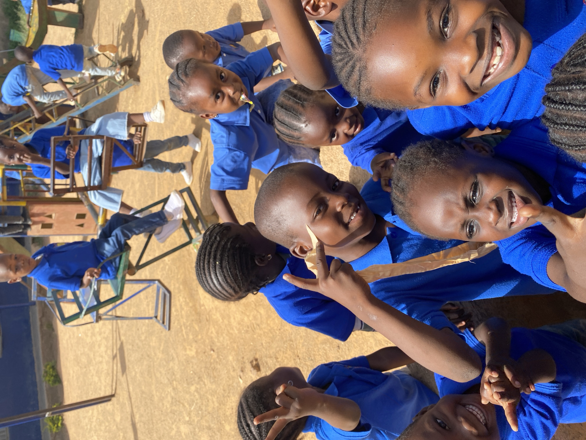 A group of children, possibly school children, are outdoors in what appears to be Gura Top, Nigeria. They are wearing blue uniforms. Some of the children are on playground swings. The ground is light brown. A group of children are taking a group selfie, smiling for the camera and making hand gestures.
