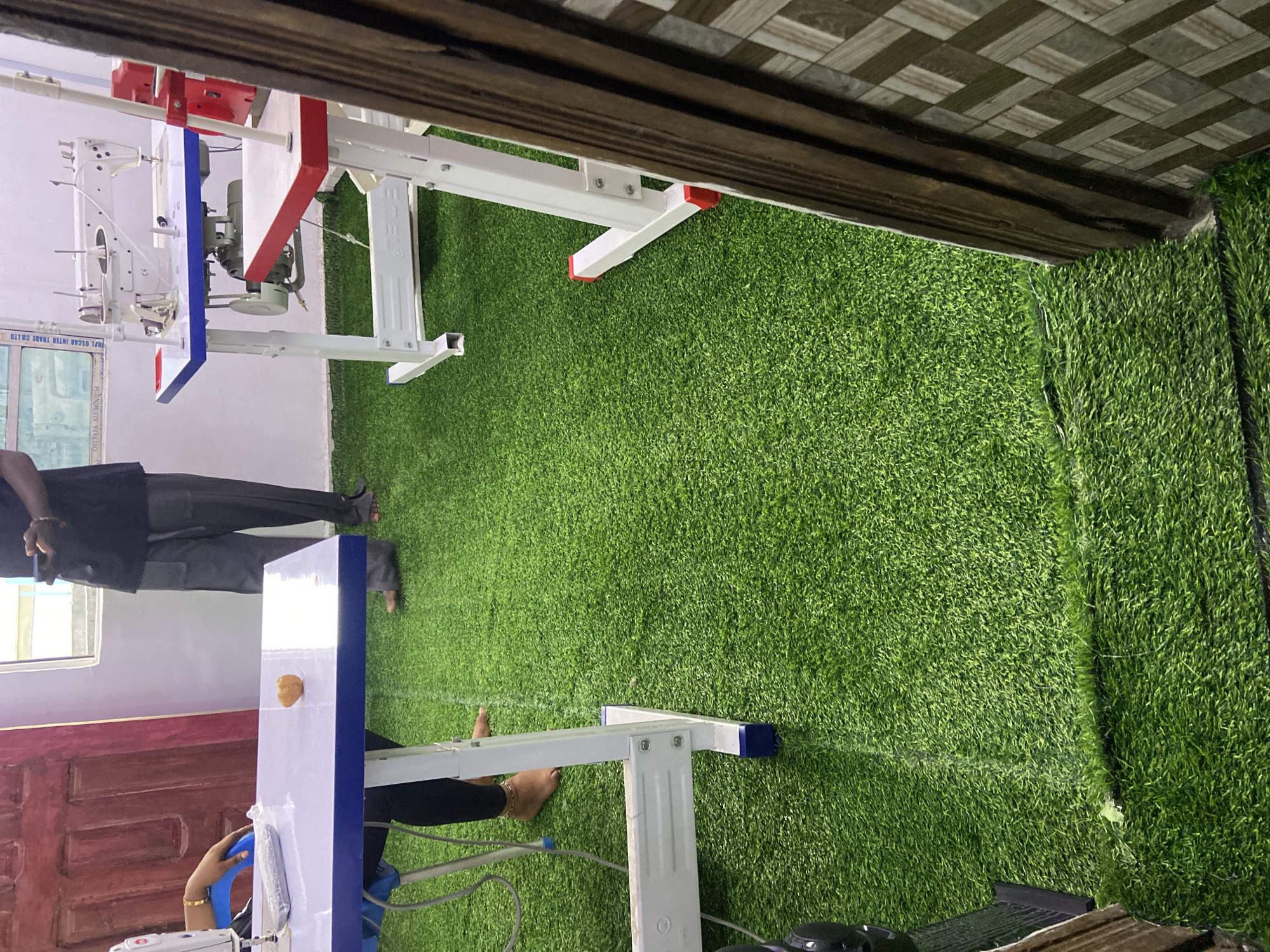 The image shows a room in Lokoja, Nigeria, with artificial turf flooring. Visible are parts of two sewing machines with white tables and metal frames with red and blue accents. An individual with dark skin wears black trousers and is partially visible. The background includes a white wall and a brown wooden door. The ceiling has a decorative brown tile pattern.