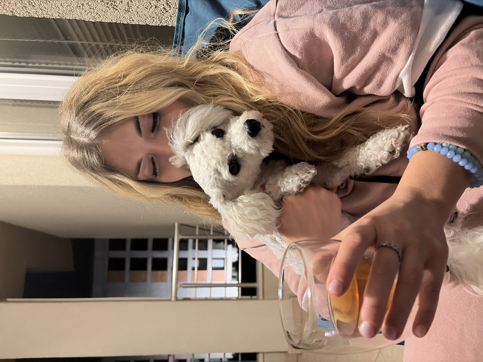 A young woman with long blonde hair, wearing a pink hoodie, is holding a small, white dog against her chest. She is also holding a glass containing a light-colored liquid. Her eyes are closed, and she appears to be resting her head against the dog. She is wearing a blue beaded bracelet on her wrist. The setting seems to be a balcony in an apartment building, as indicated by the windows and railings visible in the background. Based on the architectural style visible in the background and the colors, the location appears to be in Budva, Montenegro.