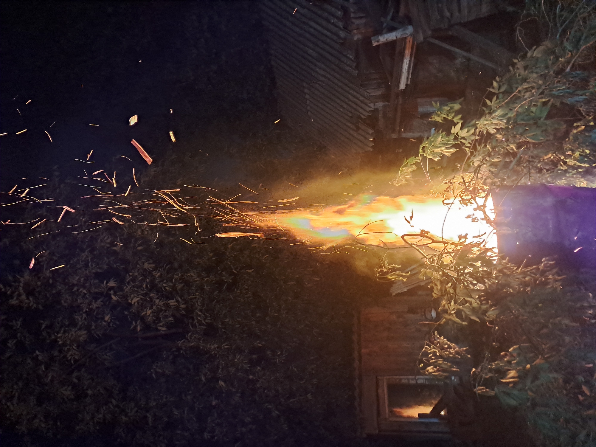 An outdoor, vertically oriented nighttime photograph depicts a powerful stream of fire and sparks emanating from the right side of the frame, extending leftwards against a dark background. The scene is set in Polyaniki, Russia.
The primary subject is a dynamic, elongated flame, primarily orange and yellow in color, with hints of green and purple fringes, suggesting intense heat or chemical additives. Numerous bright, incandescent sparks are seen flying from the flame, appearing as long, streaking lines due to their rapid motion or photographic exposure.
To the right of the flame, dense, dark green foliage, possibly from a tree or large bush, is partially illuminated by the fire's glow. Behind the foliage and further to the right, a structure with corrugated siding, possibly a shed or part of a building, is visible, also receiving light from the blaze. In the lower right, a window reflects the bright orange light of the fire. A distinct purple glow emanates from the far right edge of the image, close to the fire's origin, which might be another light source or a unique reflection.
The dark backdrop of the night sky, visible on the left and upper parts of the image, contrasts sharply with the vivid intensity of the fire. Additional dark foliage is present in the lower left foreground. There are no visible people or discernible text in the image. The overall impression is one of a controlled but dramatic burning event in a rural or semi-rural setting.