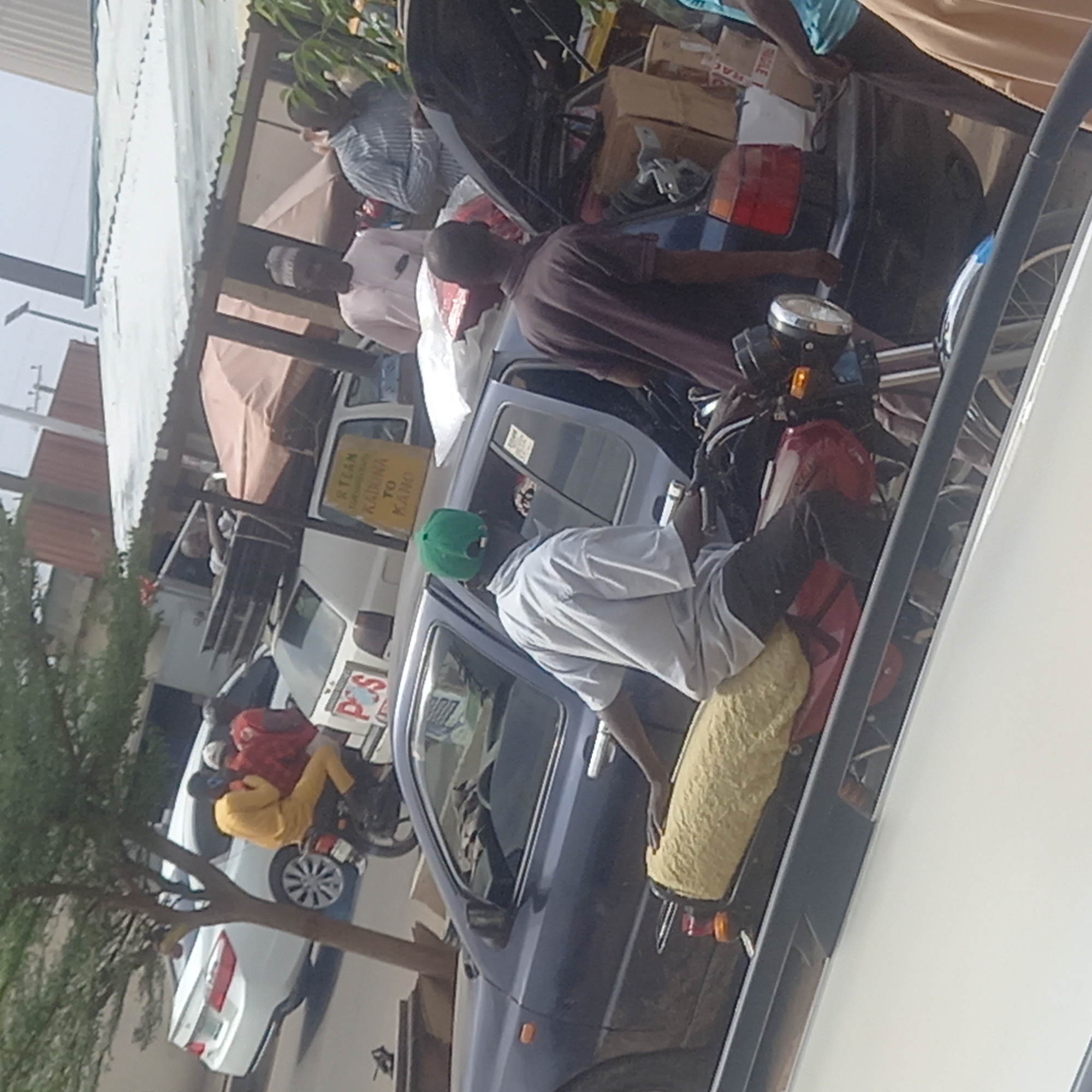 This daytime image captures a busy street scene, likely a loading or transport point in Dadi Riba, Nigeria. Several individuals are gathered around vehicles, suggesting a hub for travel or goods transfer.
In the foreground, a dark blue car has its trunk open, revealing numerous cardboard boxes and other items, indicating it is either being loaded or unloaded. Several men are positioned around this car. One man, wearing a green cap and a white long-sleeved shirt, is seated on a red motorcycle with a yellow, possibly padded, seat. He appears to be observing the activity around the blue car. Another man, dressed in a brown polo shirt, stands next to the blue car, facing away from the viewer. Further to the left, another man in a light-colored striped shirt is visible near the blue car's rear.
In the midground, a white car is parked. Next to it stands a yellow sign with black lettering. Visible text on this sign reads "RTEAN" (partially visible at the top) and clearly "KADUNA TO KANO," indicating a transport route between these Nigerian cities. Below the "KANO" text, there's another word partially visible, possibly "SERVICE" or similar. Further back, another person in a yellow top is seated on a red and black motorcycle next to the white car. A sticker with the text "P.O.S" (Point of Sale) is visible on this motorcycle or a nearby object.
The background features a building with a corrugated metal roof, suggesting local architecture, and some green foliage from trees. The bright lighting indicates clear weather during the day. The scene conveys a sense of everyday activity and commerce related to transportation.