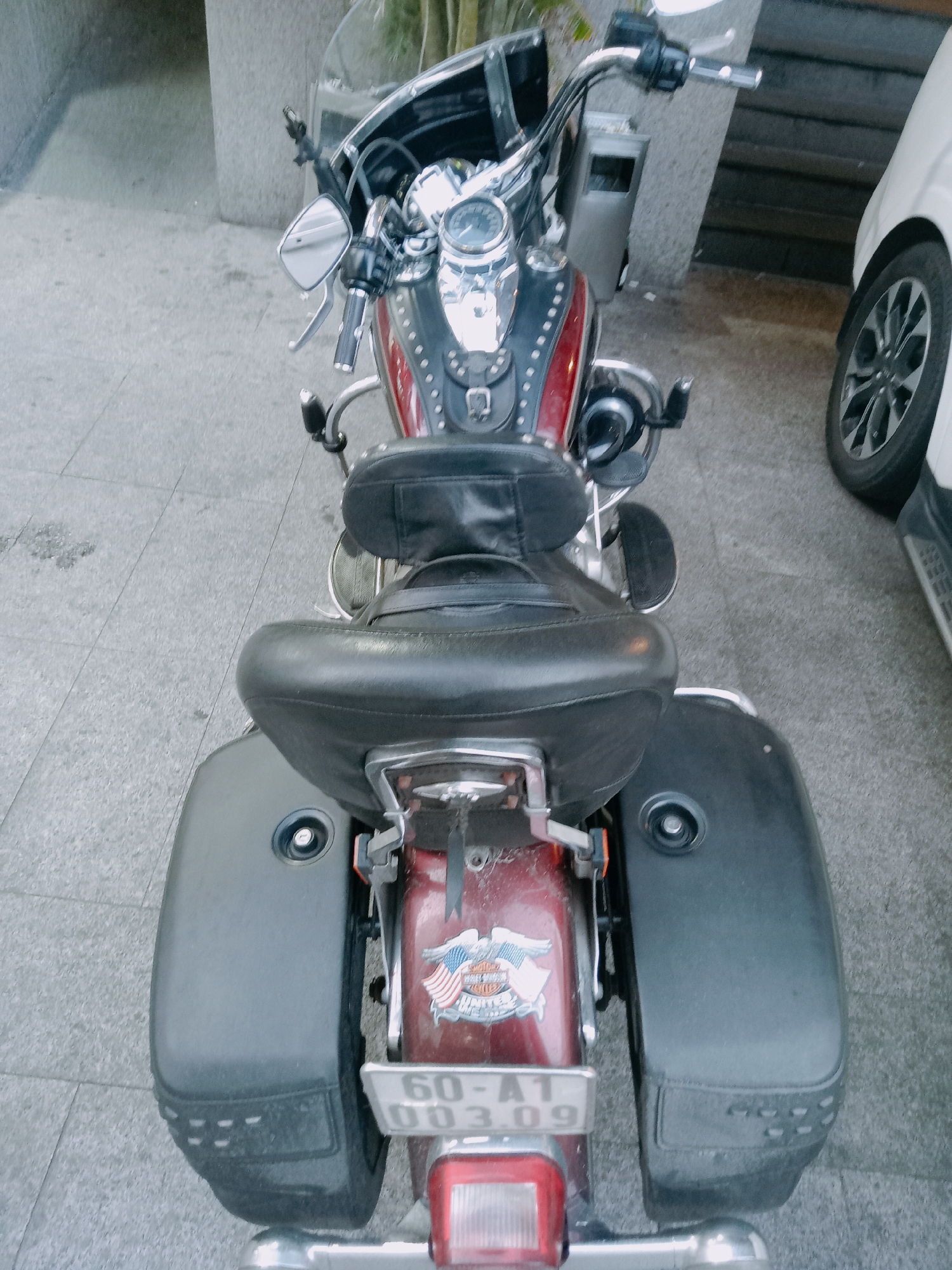 The image shows a parked motorcycle, viewed from the rear. It has saddlebags and a visible license plate. The location appears to be outdoors, possibly on a paved area or sidewalk, near a building. In the background, a small section of a car is visible. The motorcycle has a Harley Davidson logo. The license plate is partially legible with "60-A1 003.09" printed on it, which is indicative of a vehicle registered in Vietnam, specifically Nha Trang Ward.