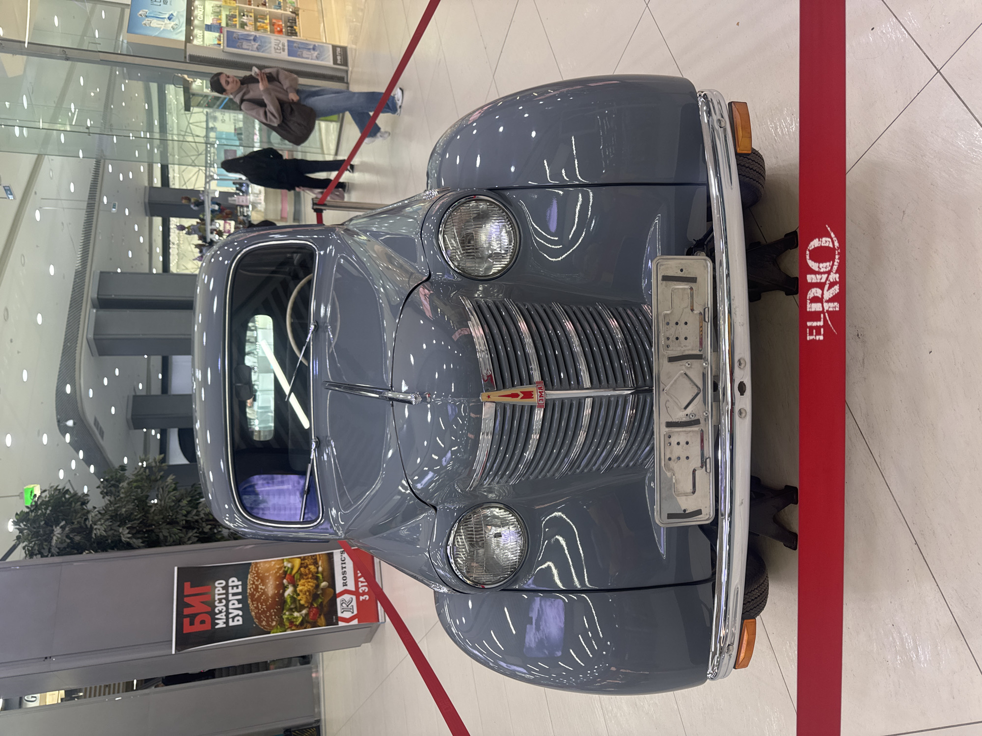 The image shows a grey vintage car, possibly a Moskvich 402, on display in a shopping mall. The car is the central focus, positioned on a tiled floor and roped off with red tape. Behind the car, there is a poster advertising a "BIG MAESTRO BURGER" from "ROSTIC'S" and indicating "3rd FLOOR". In the background, several people are visible, walking through the mall. The environment appears to be a modern indoor shopping complex with bright overhead lighting and glass storefronts. The location is identified as Samara, Russia.