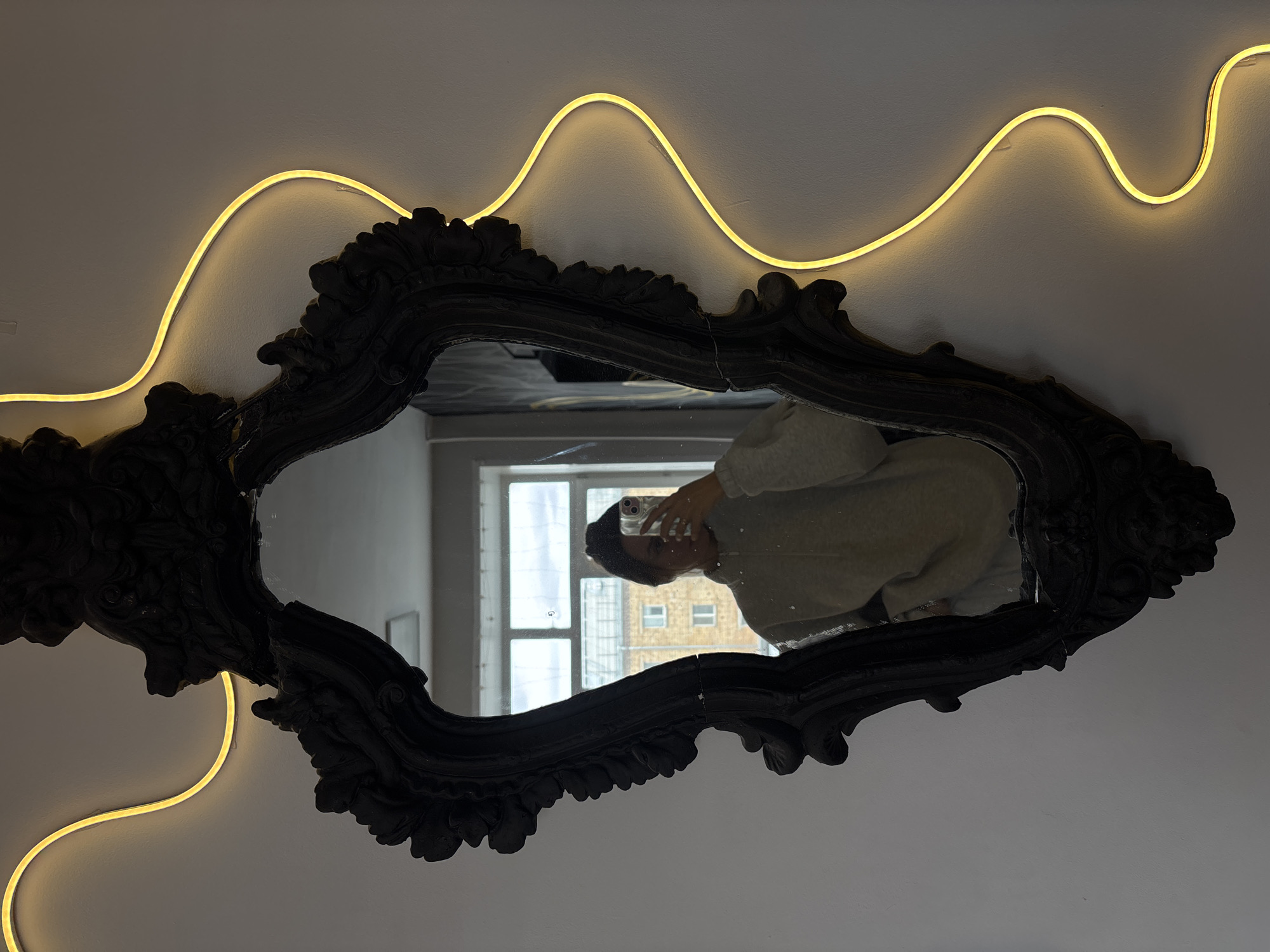 A photograph depicts a dark, ornate, baroque-style mirror, reflecting a person taking a selfie with a mobile phone. The reflection includes an exterior view through a window, suggesting a cityscape. The mirror is mounted on a white wall, flanked by a sinuous, yellow neon light fixture. The setting appears to be indoors, possibly an apartment or studio in Krasnoyarsk, Russia, based on architectural details visible in the reflected cityscape. The person in the reflection is wearing a light-colored sweater or jacket.