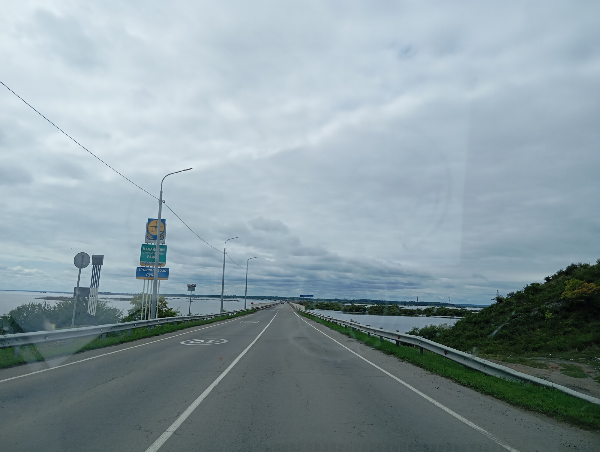 The image depicts a road leading across a bridge over a body of water. The sky is overcast with gray clouds. On the left side of the road, there are signs, including a yellow sign with a picture of a man and text, and a blue sign with text. A street lamp is also visible on the left. The road has lane markings and a speed limit sign of 40. The right side of the road has a metal guardrail and a grassy embankment with trees and some debris at the bottom. The body of water appears to extend into the distance, with some islands or landmasses covered in vegetation. In the far distance, there are some industrial structures and power lines. The overall impression is of a somewhat industrial or rural setting, possibly in Russia, given the Cyrillic text on the signs. The location is identified as Mayak, Russia.