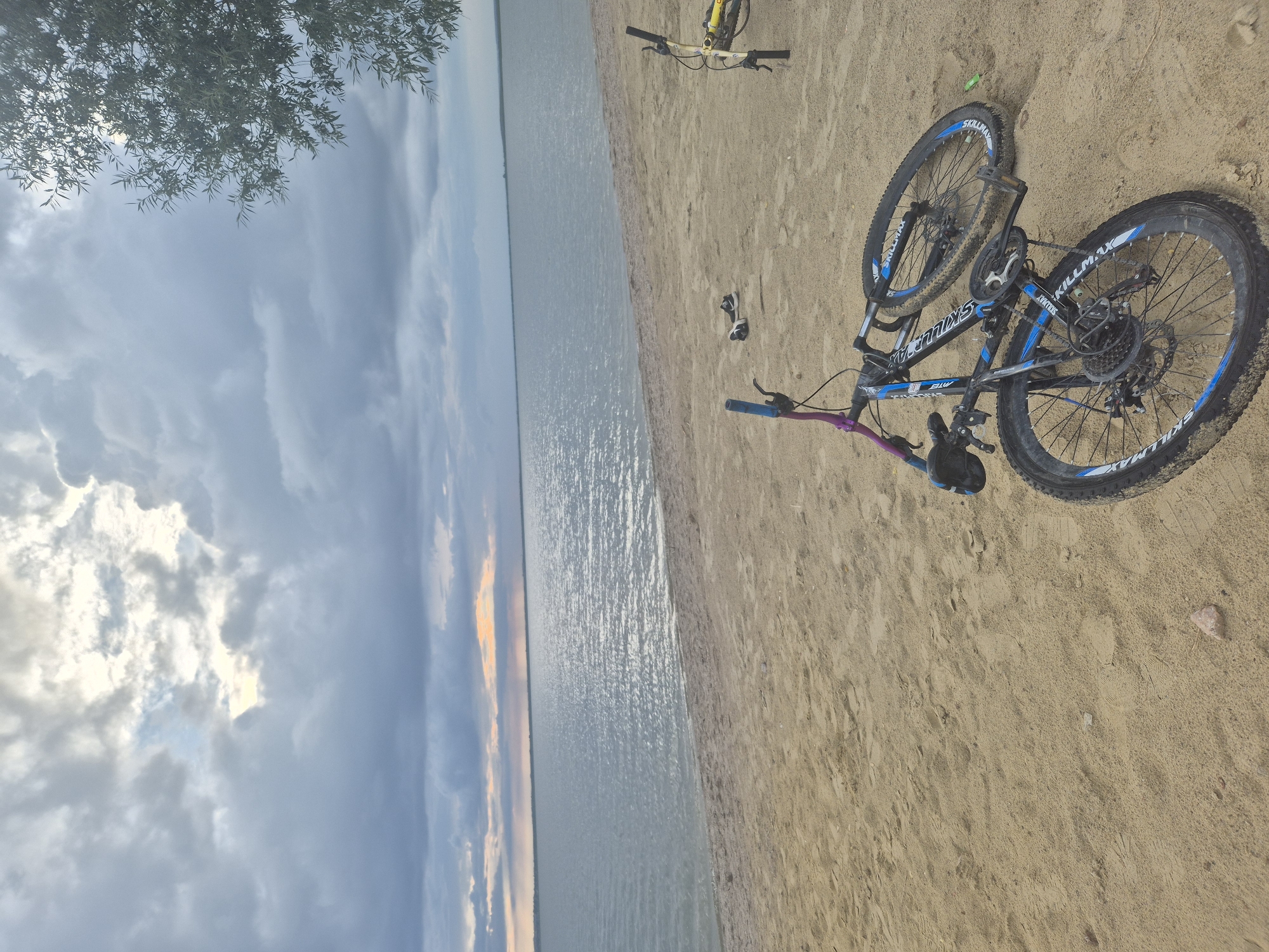 The image depicts a serene lakeside scene on a sandy beach, possibly in Kokshetau, Kazakhstan. Two bicycles are the primary subjects, with one lying on its side in the foreground, its blue and black frame contrasting with the tan sand. Its wheels are caked with mud, suggesting recent use. A second bicycle, mostly yellow and black, stands upright on the right side of the frame, its front wheel turned. A pair of black sneakers with white soles rests on the sand between the two bicycles.
The background is dominated by a vast, calm body of water that stretches to the horizon, meeting a dramatic sky filled with heavy, gray clouds interspersed with patches of bright, filtered sunlight. The clouds create a diffused lighting effect, casting a soft glow on the water's surface, which is rippled and reflects the sky. A lone tree branch, with delicate green leaves, extends into the frame from the upper right corner, its presence adding a natural element to the composition. The sand on the beach is lightly textured with footprints, hinting at previous activity. The overall atmosphere is one of quiet solitude and natural beauty.