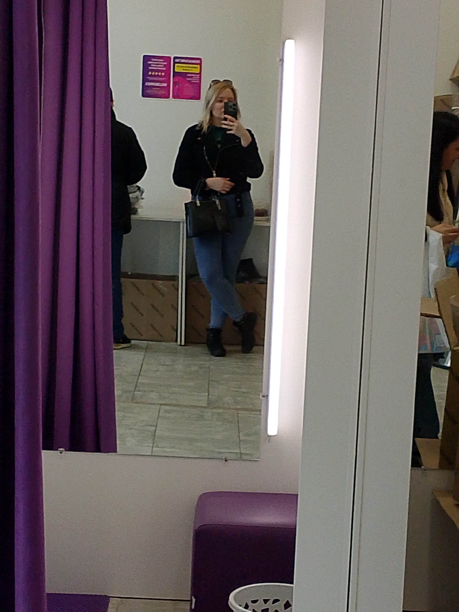 The image shows a person, presumably a woman, taking a selfie in a fitting room. She is wearing a black jacket, blue jeans, and black boots, and carrying a black purse. Her blonde hair is partially up, and sunglasses are perched on her head. The background reveals another person standing near a shelf, and a third person can be seen in the reflection of another mirror. The fitting room has purple curtains and a purple ottoman. Based on the presence of informational posters in Russian, the location is likely Izhevsk, Russia. The environment appears to be a clothing store fitting room.