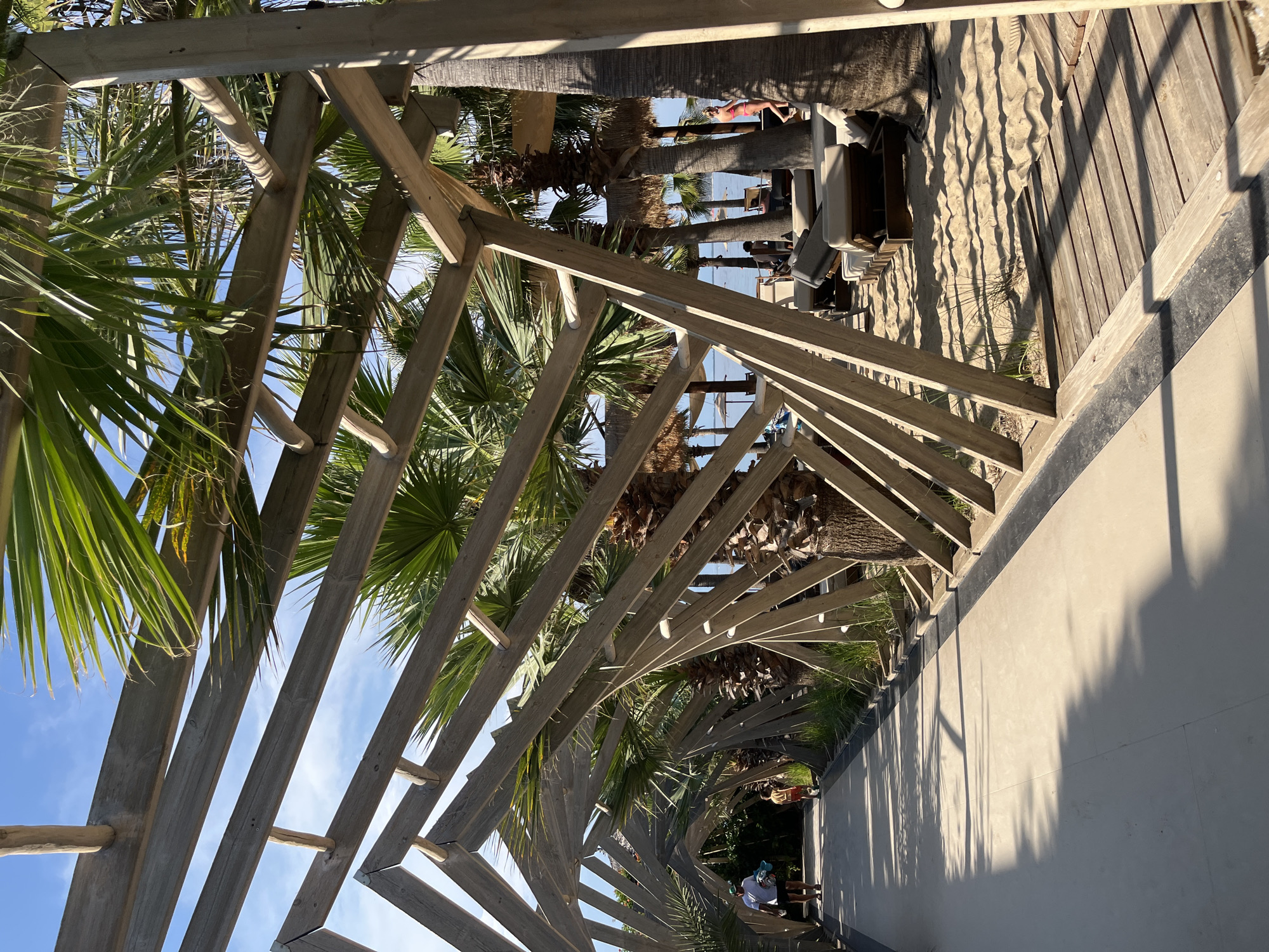 The image depicts a tropical beach scene under a clear blue sky with white clouds. A pathway runs along the left side of the frame, shaded by a wooden pergola structure made of angled beams. Palm trees with lush green fronds are visible throughout the scene, some growing within the pergola structure and others outside.
On the pathway, a man wearing a white t-shirt, dark shorts, and a hat walks away from the camera, carrying a light blue bag in his left hand. Beside him, a child in a red t-shirt and shorts walks ahead.
To the right of the pathway, the scene transitions to a sandy beach area. Several lounge chairs with beige cushions are arranged on the sand, some under umbrellas. In the background, the turquoise ocean is visible, with a few people scattered on the beach and near the water. The architecture of the pergola, with its geometric wooden design, suggests a modern beach club or resort setting. The overall atmosphere is relaxed and summery.
It is highly likely that this image was taken in Thassos, Greece, given the characteristic palm trees, clear blue skies, and the presence of a beach resort atmosphere.