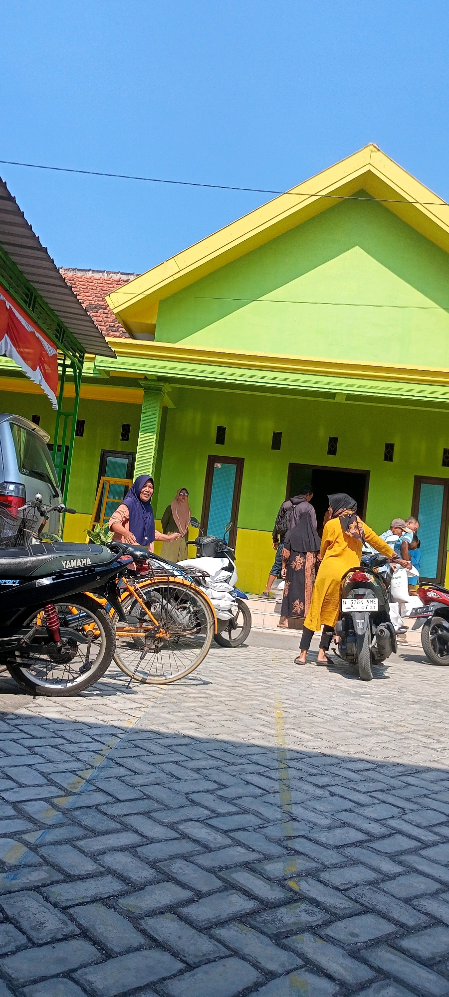 A group of people are gathered outside a bright green building with a yellow roof trim in Gempolklutuk, Indonesia. The building appears to be a public facility, possibly a school or community center, with multiple doorways. Several motorcycles and a bicycle are parked on the patterned brick pavement in front of the building, suggesting a common mode of transportation in the area. The weather is sunny with a clear blue sky.
In the foreground, a black Yamaha motorcycle is visible. To its right, an orange bicycle is parked next to a white motorcycle. Further back, a woman in a blue hijab and light brown top is standing and smiling. Behind her, another woman in a light brown hijab is near a motorcycle.
To the right of the building entrance, a woman in a yellow dress and black hijab is sitting on a black motorcycle, looking back. Another person in a black and floral patterned dress and black hijab is standing behind her. A man in a grey jacket is also present. A person in a light blue shirt and a white cap is visible further to the right, near a white bag. The overall atmosphere seems casual and everyday.
