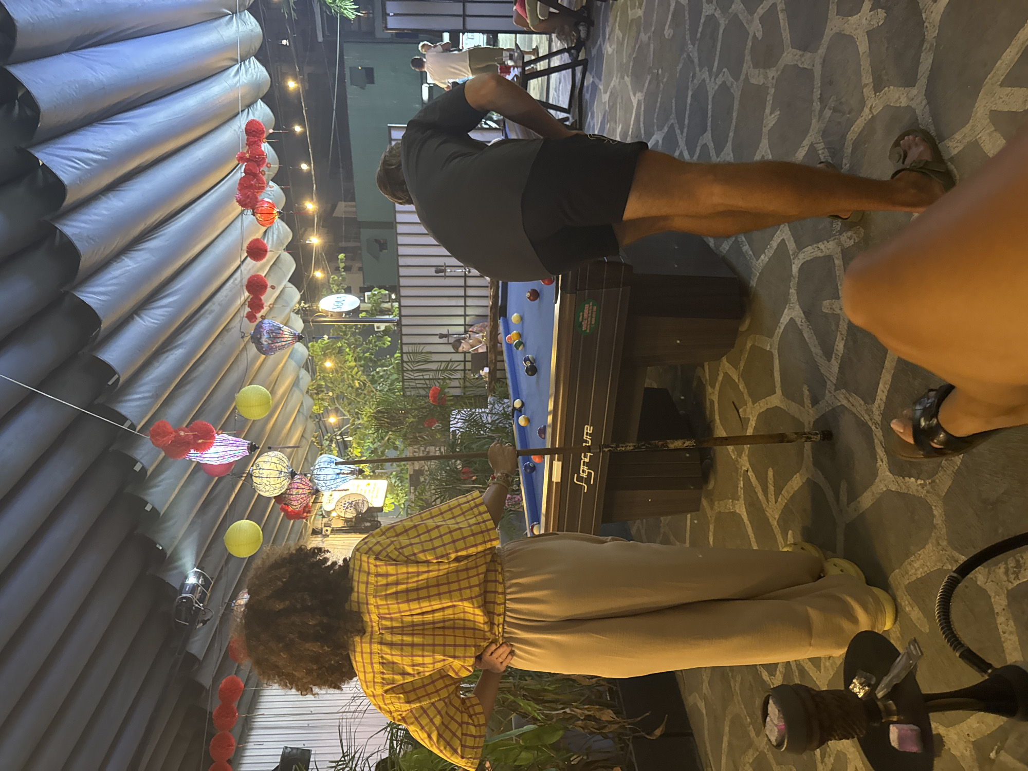 Two individuals are playing pool at night. A person in a yellow plaid shirt is positioned to take a shot at the billiard table. Another person in a dark shirt is leaning over the table. A pool cue rests on the table. Decorative lights hang above. In the foreground to the right, a hookah is visible. The location is in Đà Nẵng, Vietnam.