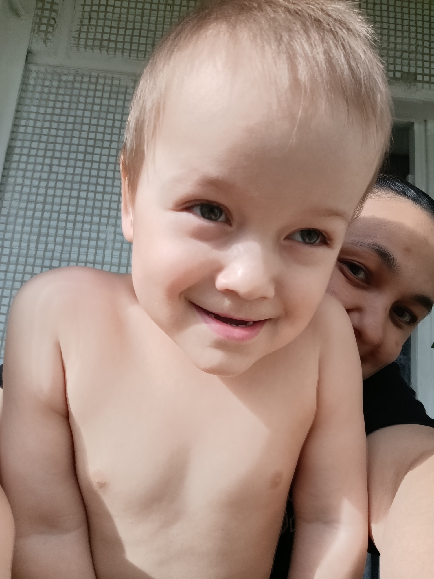 A close-up shot shows a fair-skinned, shirtless toddler smiling at the camera, held by an adult. The adult has dark hair pulled back, dark eyes, and is wearing a black shirt. The background features a light-colored tile wall. The location is identified as Chaikovsky, Russia.