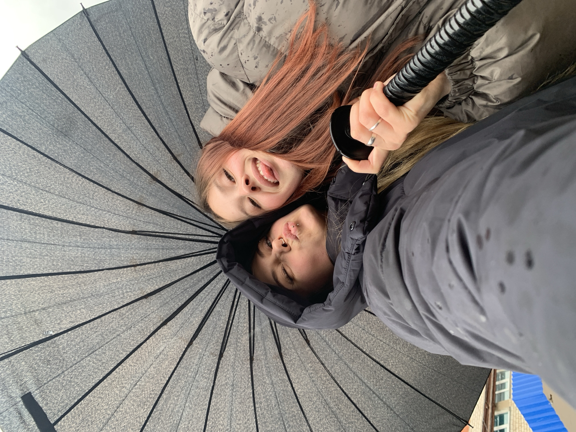 Two young women pose under an open grey umbrella, likely held up by one of them, indicated by a hand visible near the handle. The woman on the left has red hair, a light skin tone, and is sticking out her tongue with her eyes closed. The woman on the right has brown hair under the hood of a dark grey jacket. The location may be Фоки, Russia. A corner of a building with a blue roof is seen in the background. Both women are wearing winter coats, suggesting a cold or rainy season. The photograph is taken from a low-angle, looking up at the women and the umbrella.