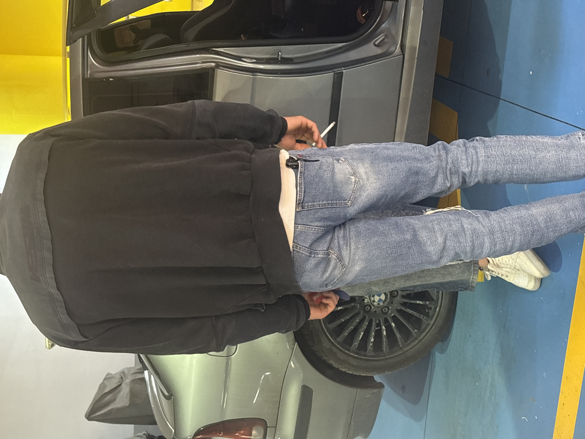A person stands facing away from the camera, wearing a black jacket, a white t-shirt, and ripped blue jeans. They are holding a lit cigarette in their right hand and have a blue object tucked into their back pocket. The person is standing next to a silver car with an open door, and a portion of a tire with a BMW logo is visible. The ground is blue with yellow lines. The background is a concrete wall and a yellow wall. The lighting suggests an indoor environment, possibly a parking garage.