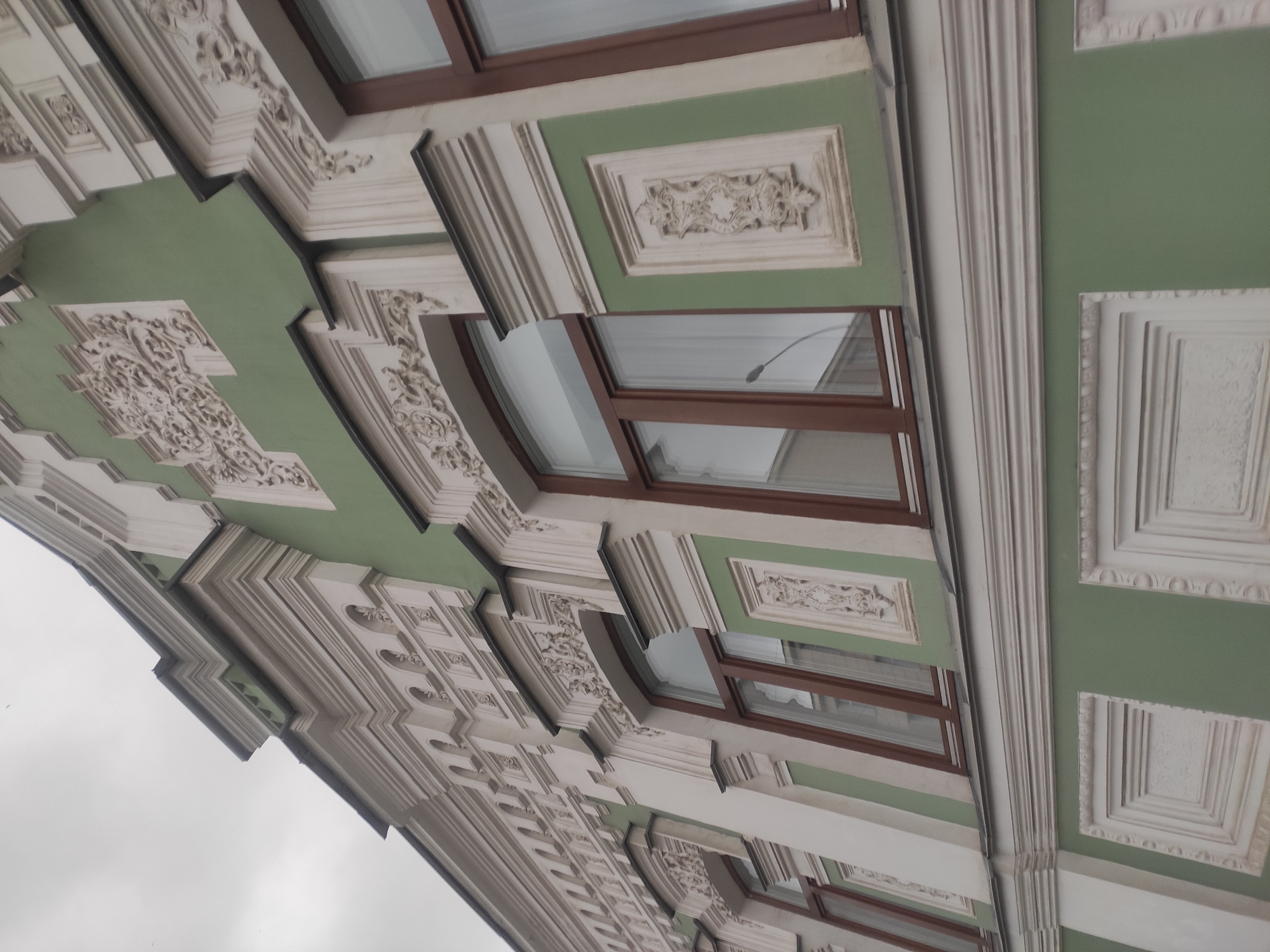 The image shows an ornate building facade in Yekaterinburg, Russia. The architecture features intricate white stucco decorations against a light green painted background. The building has several windows with brown frames. The style suggests it could be a historic building, possibly from the late 19th or early 20th century. The sky is overcast, indicating a cloudy day. There are no people or vehicles visible in the frame. The perspective is from below, looking up at the building.