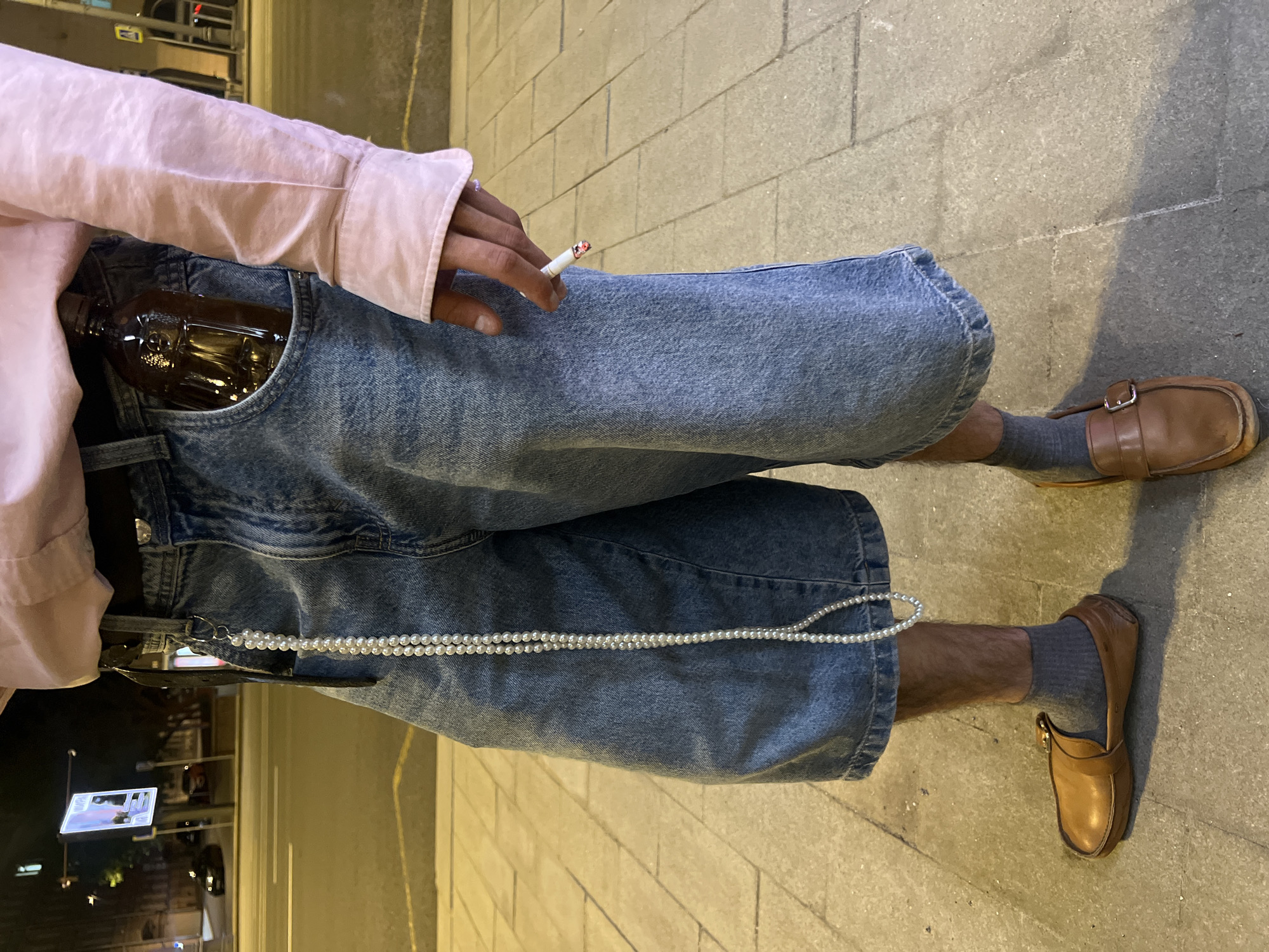 A person stands outdoors on a paved surface that suggests a sidewalk or plaza. The individual is dressed in light-wash denim shorts, brown leather loafers with grey socks, and a pink shirt. They hold a lit cigarette in their hand. A dark-colored bottle is visible in their front pocket. A string of white beads hangs from their shorts. The setting appears to be urban, indicated by the streetlights, buildings, and traffic visible in the background. The location is not recognizable as Moscow, Russia based on the visual information provided.