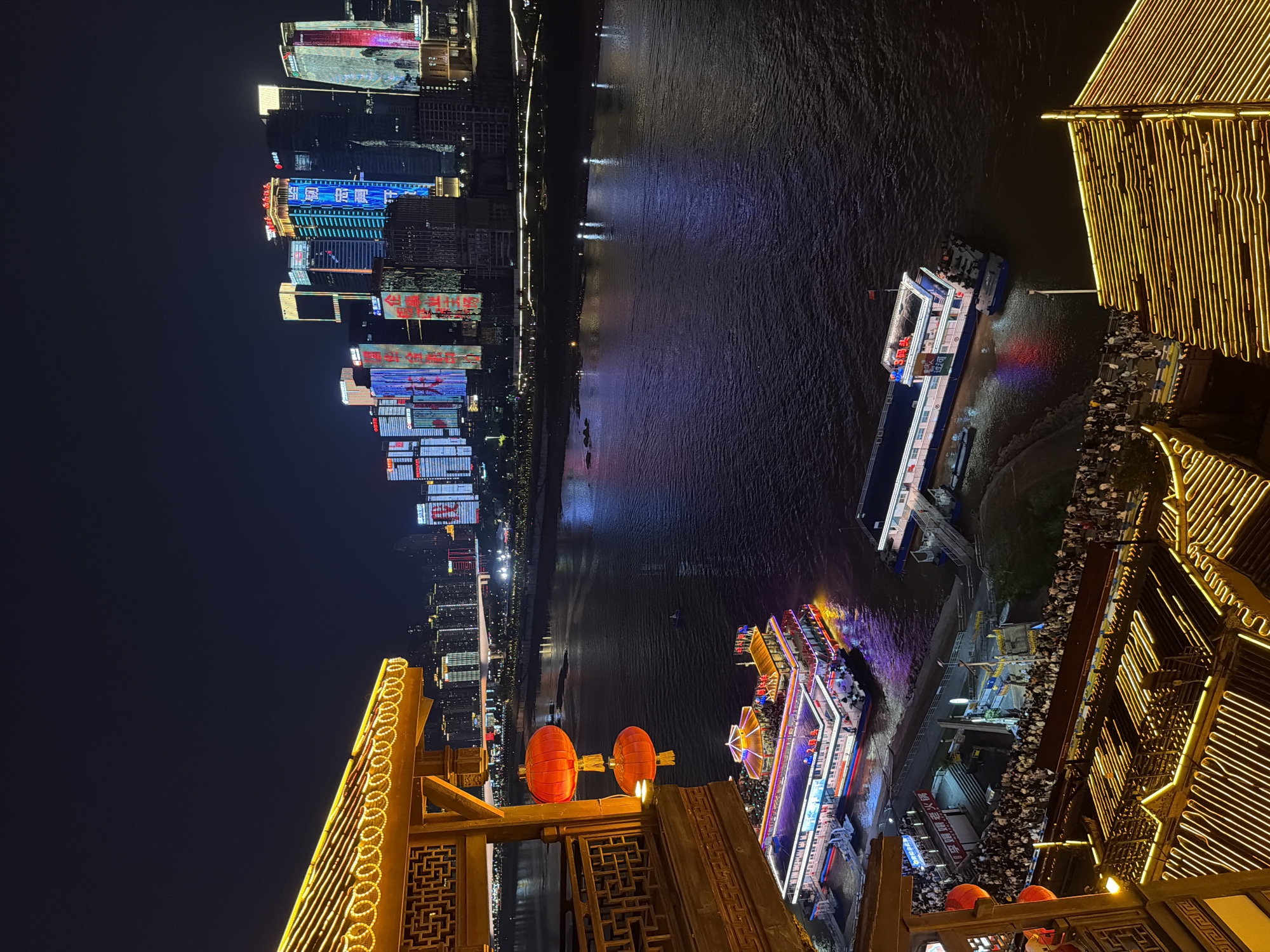 This is a night-time, high-angle, long shot of a riverfront scene in Yuzhong District, China. The foreground features traditional Chinese architecture with illuminated yellow lights along the roofs and eaves. Two red lanterns hang from a wooden balcony on the left. In the middle ground, a large, multi-story tour boat, illuminated with colorful lights, is docked. Many people are visible on the boat's decks and disembarking onto a pier. Another smaller boat is docked beside it. A large crowd of people is gathered on the waterfront below the traditional architecture, near the pier. Across the wide, dark river, the background is dominated by a modern city skyline of numerous skyscrapers. These buildings are brightly lit with colorful displays and advertisements. A bridge spans the river in the distance. The water reflects the city lights and the lights of the docked boats.