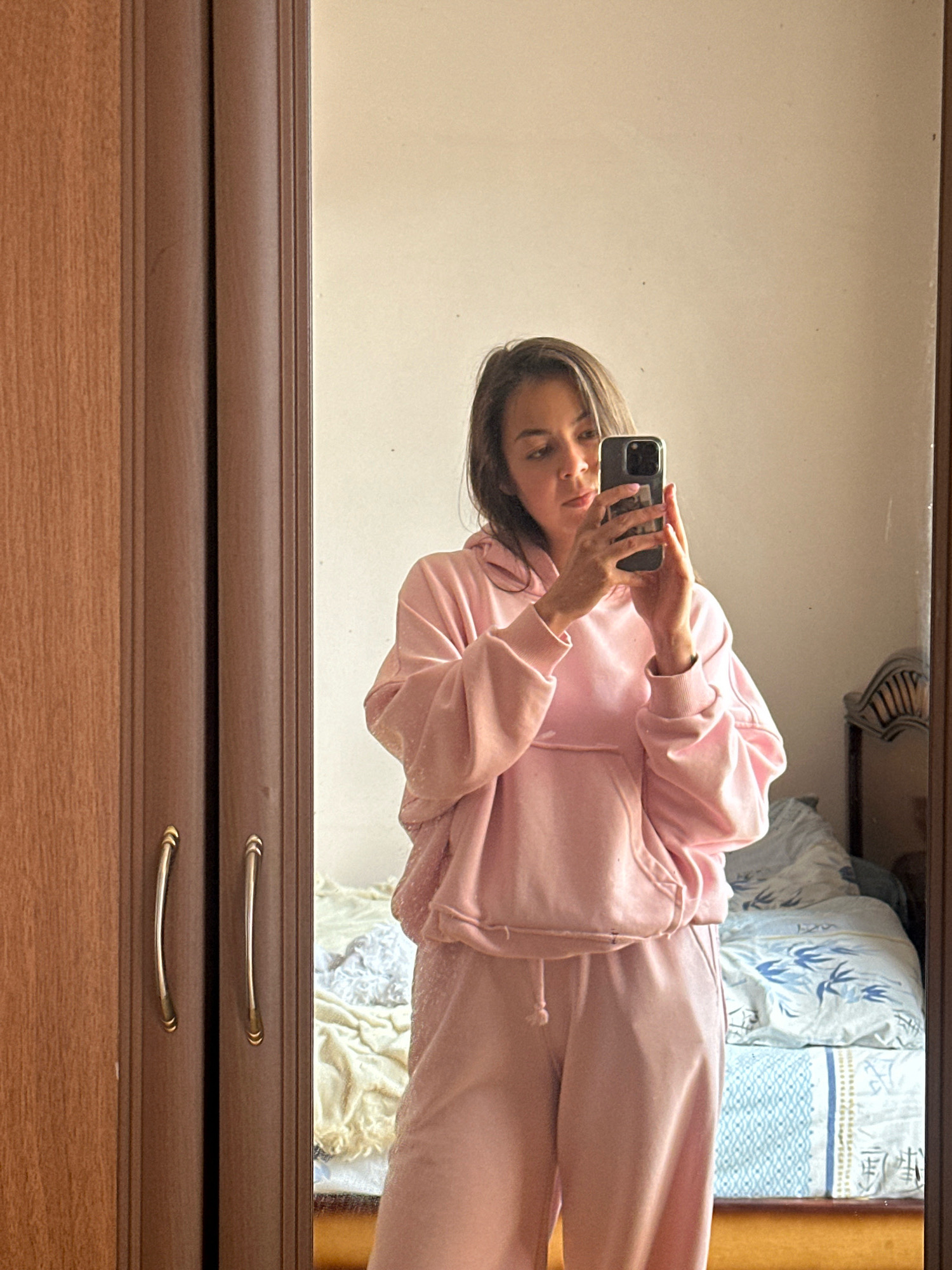 A young woman stands in front of a full-length mirror, taking a selfie. She is wearing a pink tracksuit consisting of a hooded sweatshirt with a front pocket and matching sweatpants. The tracksuit appears to be made of a soft, possibly velour or terry cloth material. The woman has brown hair styled in loose waves, and she is holding a smartphone in her right hand to capture the reflection.
The mirror is framed by a wooden wardrobe or closet, visible on the left side of the image. The wardrobe has two doors with curved, gold-colored handles. The background reflected in the mirror shows a bedroom. A bed with patterned blue and white bedding is visible, along with a portion of what appears to be a headboard with an ornate design. The walls of the room are a light, neutral color.
The image was taken indoors, likely in a residential setting. The lighting is soft and natural, suggesting it might be daytime. The overall scene is casual and personal, typical of a mirror selfie taken at home. The city of Gyumri, Armenia, is not directly indicated by any specific landmarks or environmental cues within the image itself.
