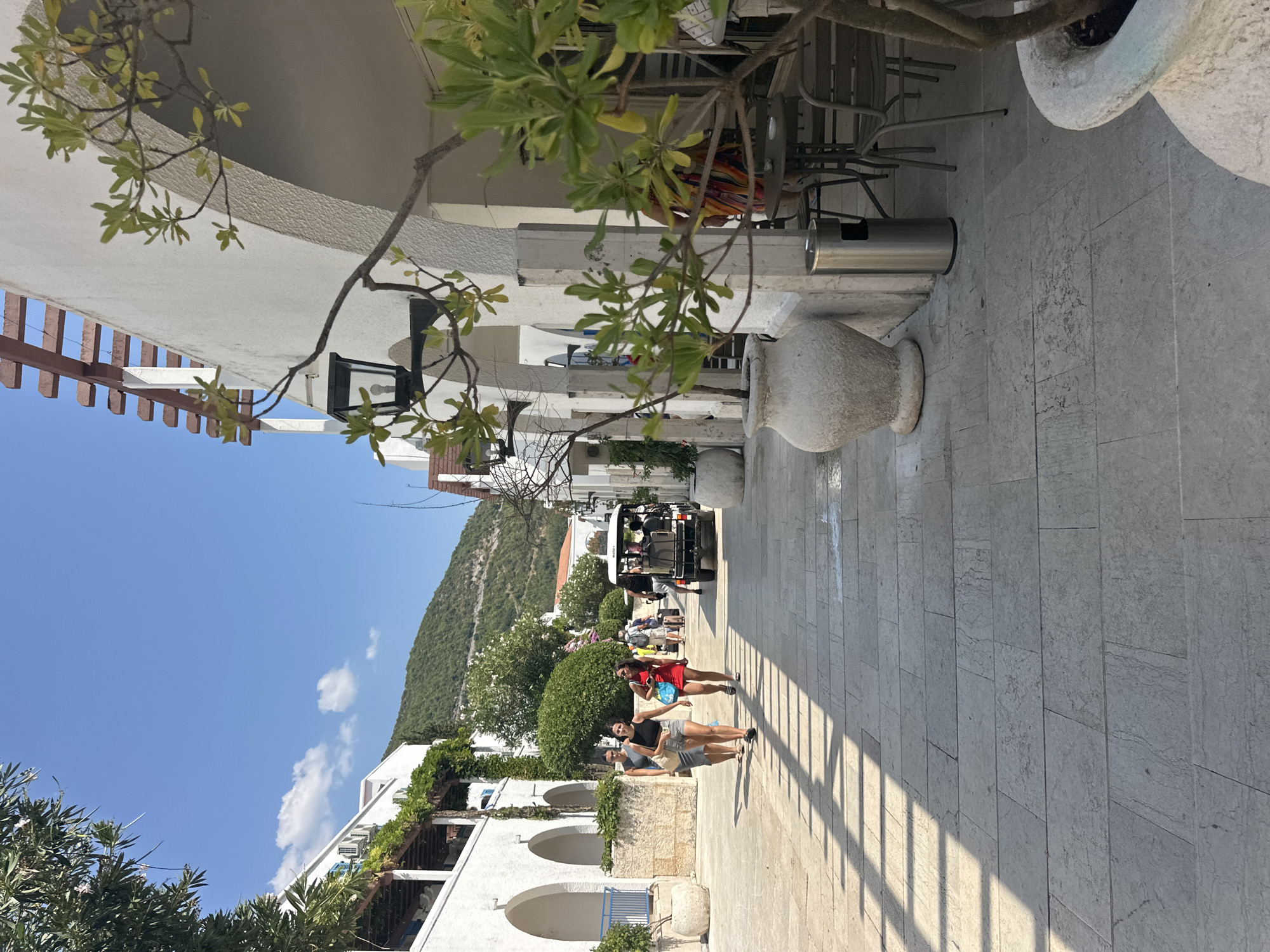 In Budva, Montenegro, a sunlit pedestrian walkway is lined with white stucco buildings featuring arched entrances and wooden pergolas. A group of people walks away from the camera down the stone-paved path, casting shadows from a nearby structure. To the right, a golf cart with passengers is visible, and further down the street, other pedestrians and a few potted plants are present. The scene is set against a backdrop of a lush green hillside under a clear blue sky.