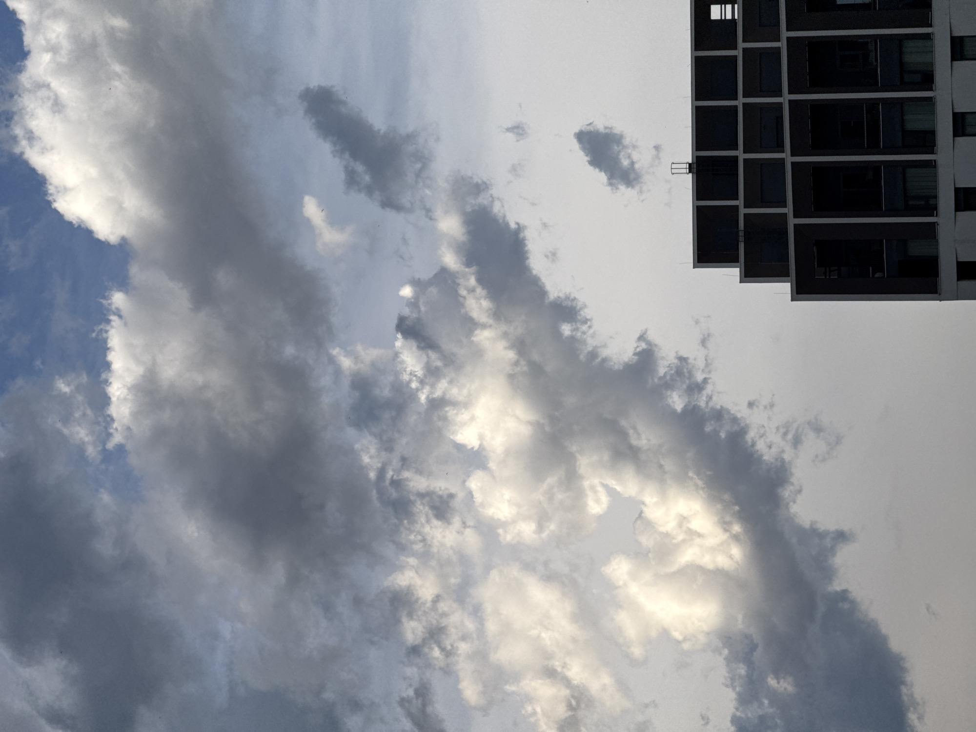 The image captures a modern, stepped building against a dramatic sky filled with dynamic clouds. The building, with its dark facade and repeating geometric window frames, is positioned in the lower right quadrant of the frame. Its structure suggests a residential or commercial complex. The sky above is a mix of dark, heavy clouds and brighter, illuminated areas, indicating a possibly overcast or transitional weather condition. The overall impression is one of urban architecture meeting atmospheric conditions. The specific location in Belgrade, Serbia, is not visually identifiable from the provided image.