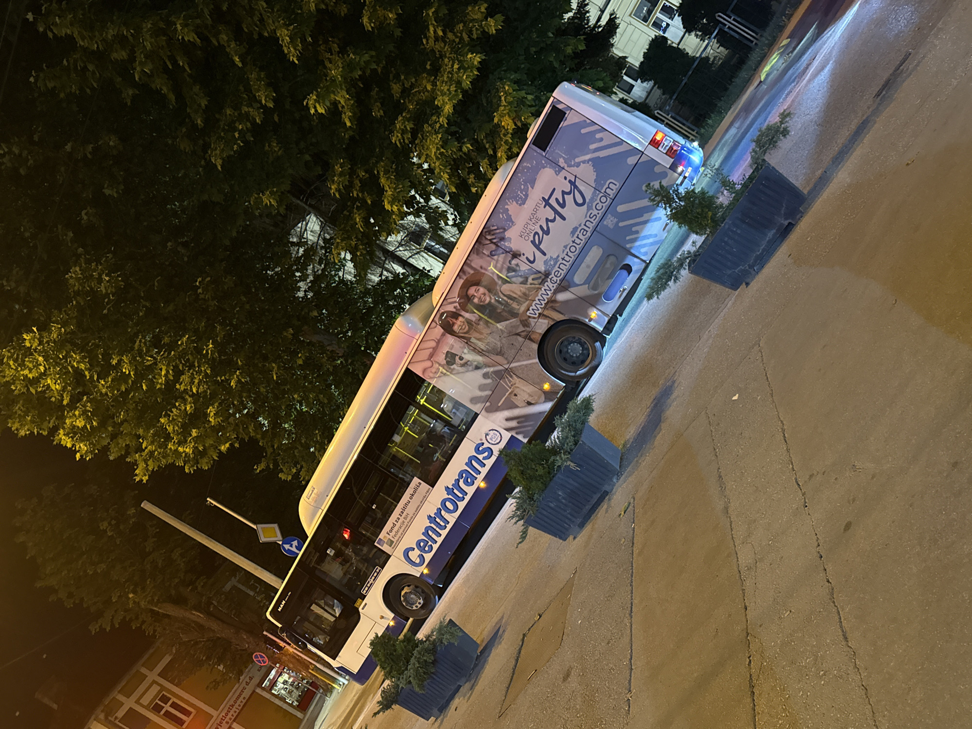 An external view of a white and blue "Centrotrans" bus parked at night on a paved street in Ilidža, Bosnia and Herzegovina. The bus features advertisements on its side, including images of people and the website "www.cent rotrans.com". The street is illuminated by streetlights, casting reflections on the wet pavement. There are planters with greenery along the sidewalk. In the background, trees and buildings are visible.