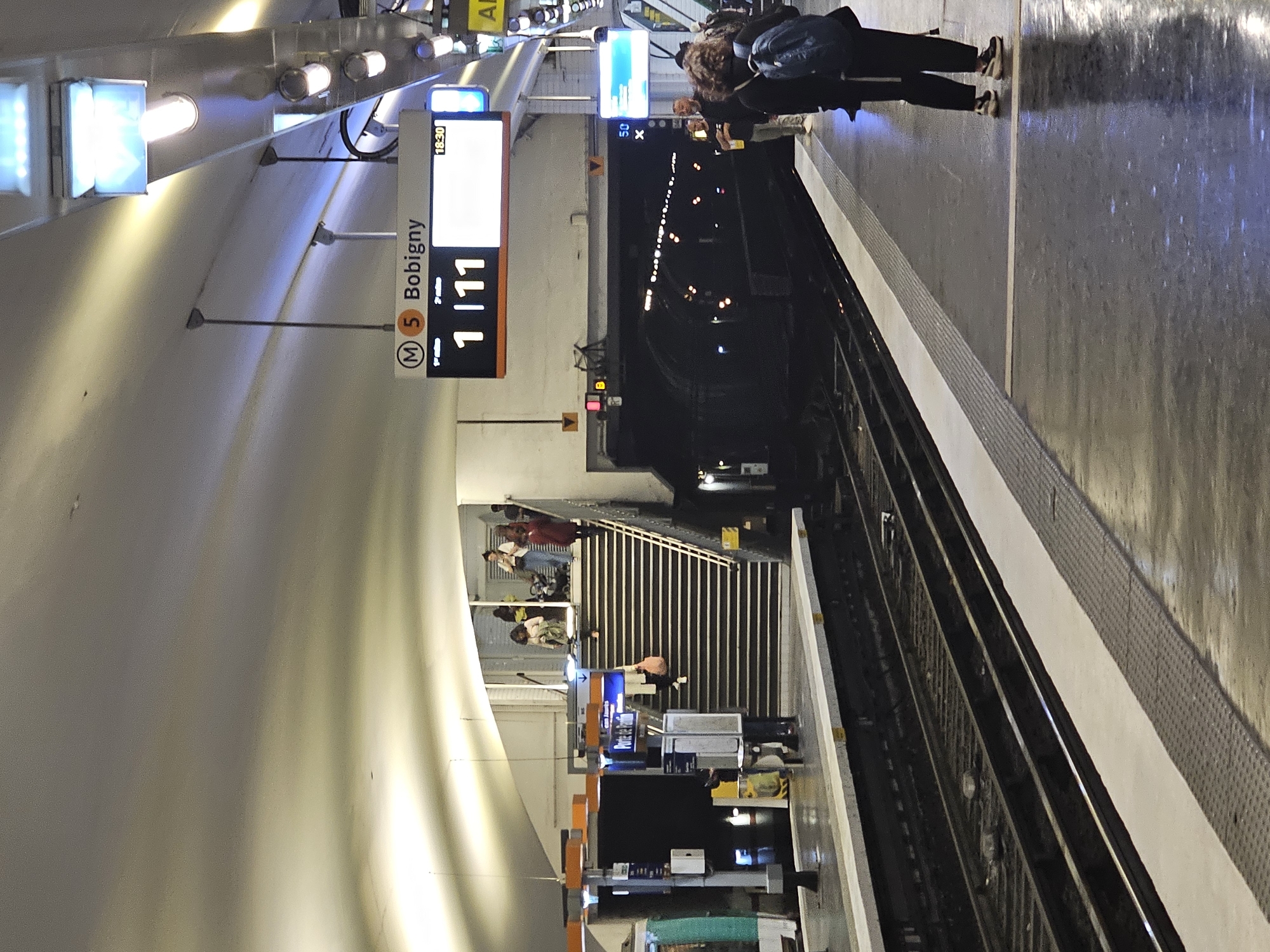 The image captures the platform of a metro station in Paris, France, specifically at the Bobigny-Pablo Picasso station on Line 5. The platform is elevated, with a view down the tracks into a dimly lit tunnel. People are standing and waiting for the train. A sign indicates the station name "Bobigny" and the upcoming train schedule, displaying "1/11" (presumably 1 minute, 11 seconds). The station's design features curved walls and illuminated signage. A staircase leading to an upper level is visible, with people ascending and descending. The overall atmosphere is that of a typical Parisian metro station scene.