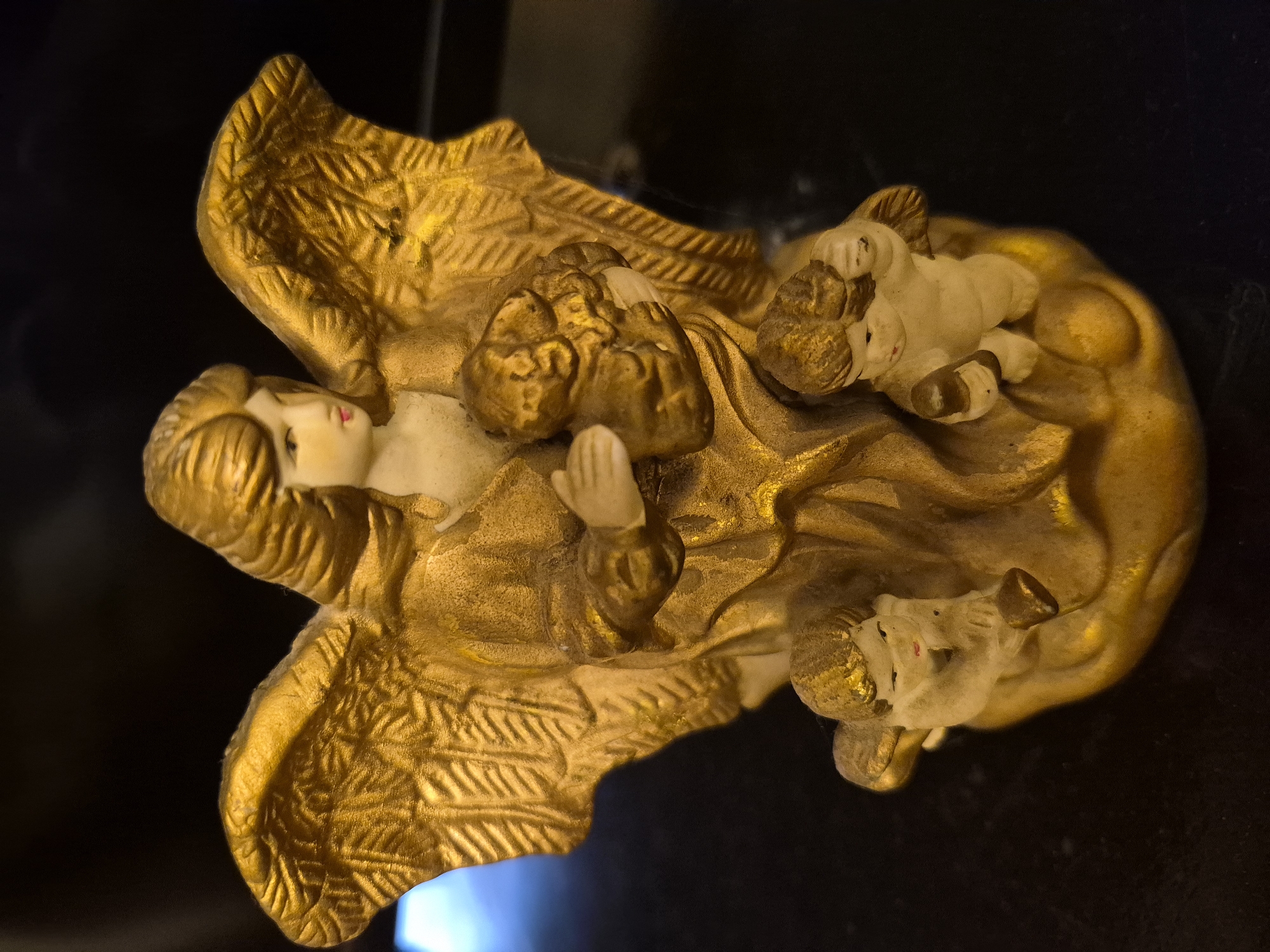 A gilded angel figurine is the subject of this image. The angel has golden wings and is holding a small golden dog. Two smaller cherubic figures with golden hair are positioned at the base of the angel. The figurine is set against a dark background. There is no indication of its location in the city of Vanadzor, Armenia.