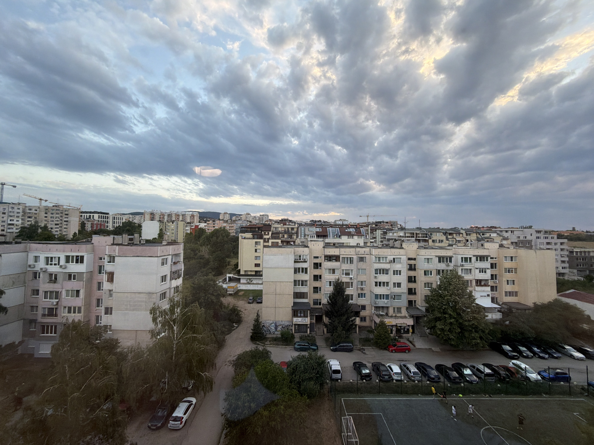 The image depicts an urban landscape in Sofia, Bulgaria, under a cloudy sky. The scene is dominated by residential apartment buildings, typical of Soviet-era architecture, with multiple stories and balconies. In the foreground, a street curves through the scene, with several parked cars. These include a white sedan, a dark sedan, and other dark-colored vehicles. Further into the image, a row of parked cars is visible along the side of a street, featuring a mix of sedans and SUVs in various dark colors, along with a few white cars and a blue vehicle. Adjacent to the parking area, a sports field with white markings and goalposts is present, where a few individuals appear to be playing soccer. Construction cranes are visible in the distance, suggesting ongoing development in the city. Trees, some appearing mature and others younger, are interspersed throughout the urban environment, providing a touch of greenery. The overall impression is of a typical residential neighborhood in Sofia, likely during the daytime or early evening given the light conditions.