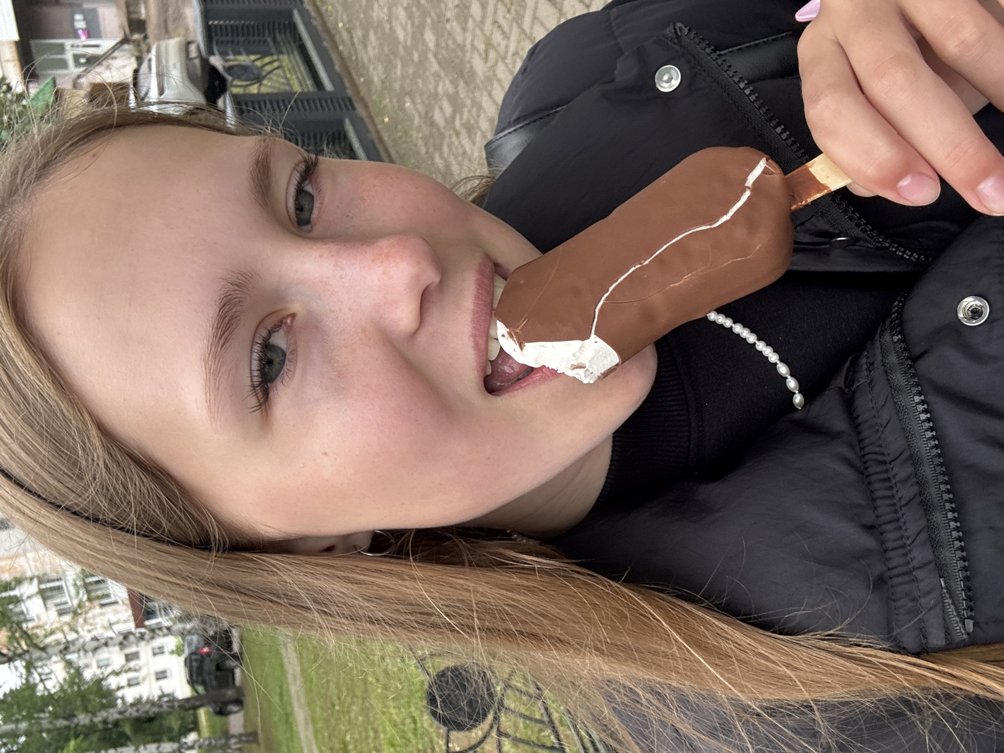 The image shows a close-up of a young girl eating an ice cream bar. She is wearing a black puffer jacket. The ice cream bar is dipped in chocolate with a white drizzle. Her face is angled towards the camera, and her mouth is open as she takes a bite. The background is blurred but appears to be an outdoor setting with some buildings and greenery visible. The lighting suggests it is daytime. The girl's expression is one of enjoyment. The image has a casual, candid feel.