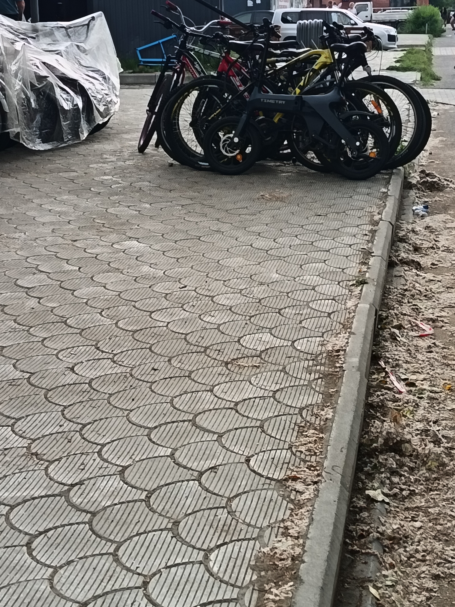 The image depicts a sidewalk with a patterned grey paving stone surface in Komsomolsk-on-Amur, Russia. Several bicycles are parked on the sidewalk. One bicycle, partially covered by a clear plastic sheet, is on the left side of the frame. Other bicycles are visible to the right, including a yellow one and a black one with the word "TIMETRY" on its frame. The bicycles are lined up, and their wheels and handlebars are visible. The sidewalk is bordered by a concrete curb, and to the right of the curb is a patch of ground covered with dry leaves and debris. In the background, a white vehicle is parked, and some vegetation can be seen. The lighting suggests it is daytime.