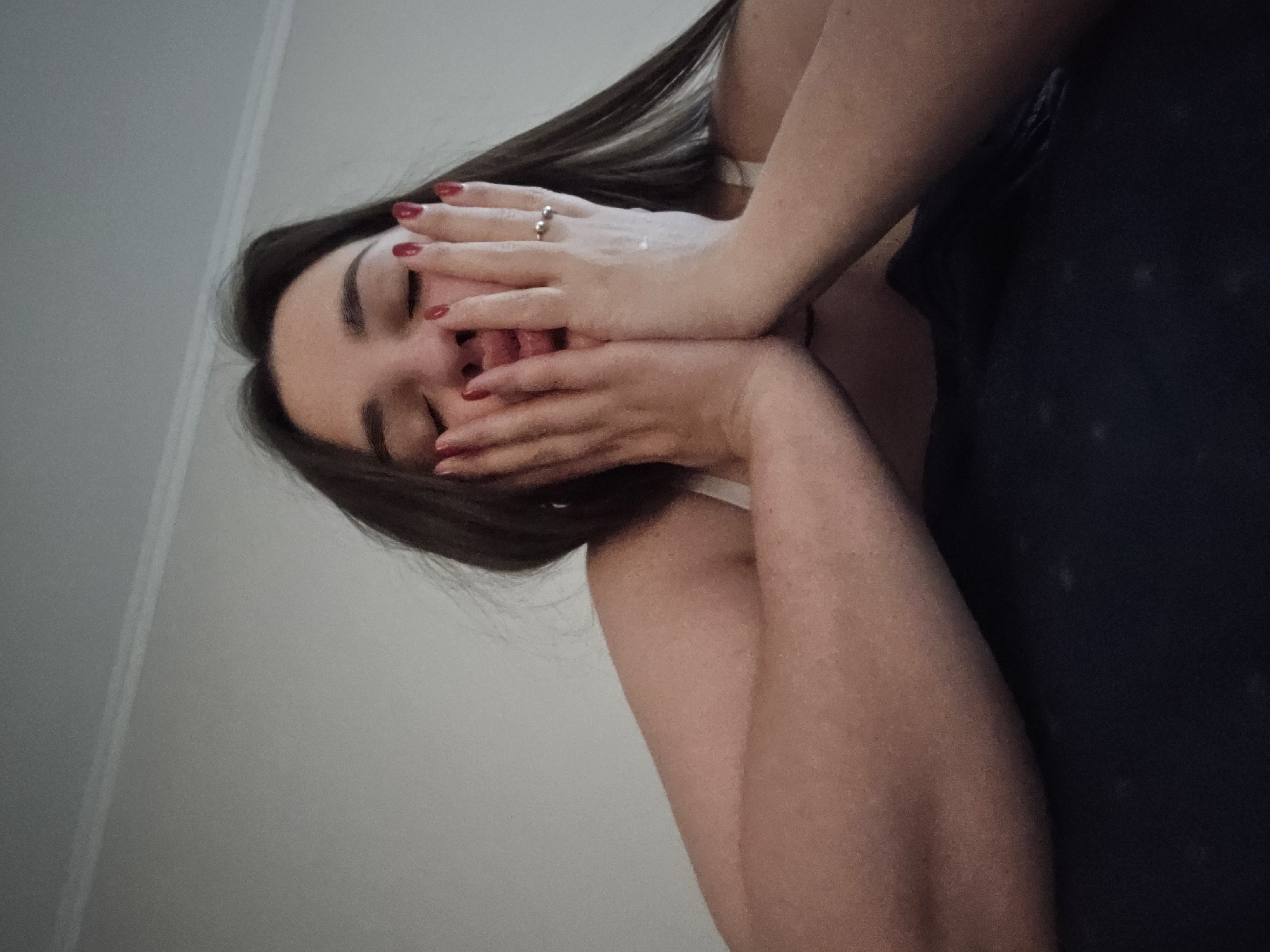 A woman is captured in a low-angle, close-up shot, her eyes closed as she rests her face in her hands. Her dark hair cascades around her shoulders, and her fingernails are painted a vibrant red. A silver ring adorns one of her fingers. She is wearing a white undergarment and appears to be under a dark blanket. The background is a plain, neutral wall. The image does not provide any visual cues to suggest a specific location in Petropavlivska Borshchahivka, Ukraine, or any specific environmental details.