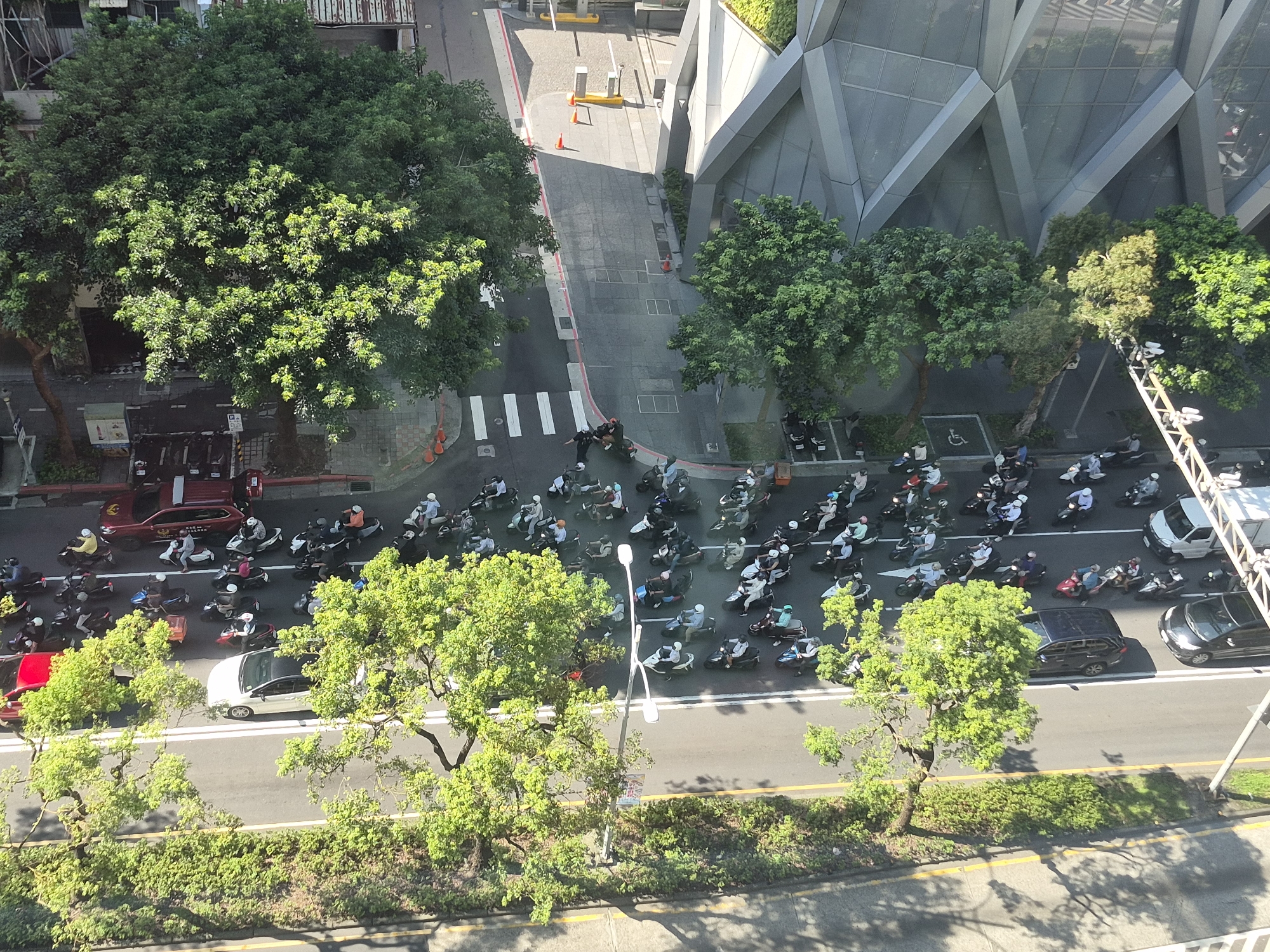 This is an aerial view of a street in Taipei, Taiwan, with a prominent modern building with a distinctive angular facade on the right. The street is lined with trees and filled with numerous motorbikes, indicating a busy urban environment. A pedestrian crossing is visible on the left side of the road, and several traffic cones are placed in the vicinity.
The scene captures a traffic flow of motorbikes, many with riders wearing helmets and some with passengers. The motorbikes are varied in color and style, typical of urban transportation in Taipei. Cars, including a red SUV and a white sedan, are also present on the road.
The building on the right displays a contemporary architectural style, with large glass panels and geometric shapes. Trees with dense green foliage line the street, casting shadows on the road, suggesting it is daytime. The overall impression is a dynamic snapshot of daily life and traffic in a bustling Taiwanese city.