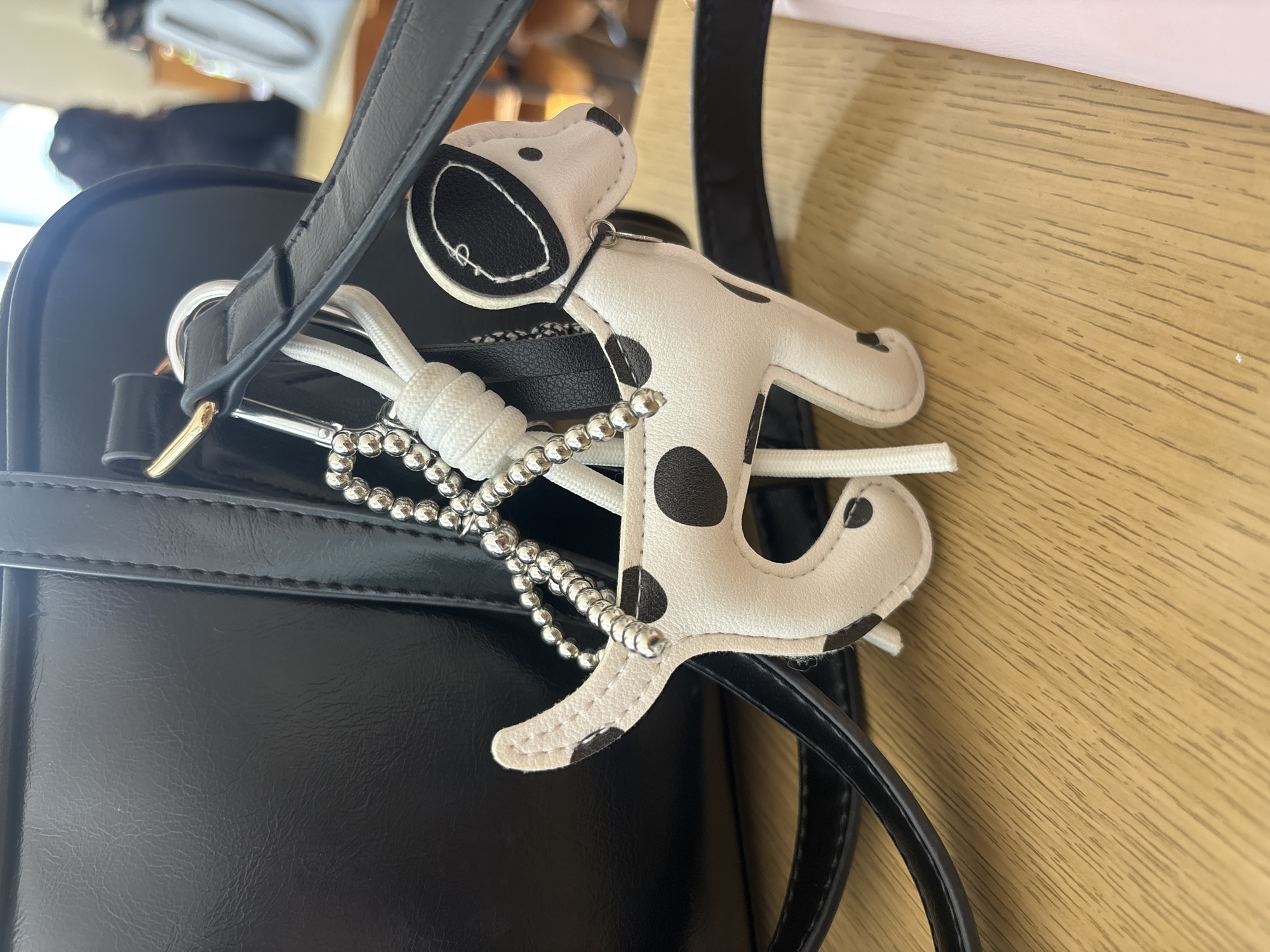 A close-up shot shows a black handbag with a dog-shaped keyring attached, positioned on a wooden surface. The keyring is white with black spots, resembling a Dalmatian, and is connected to the bag via a silver beaded chain and a white knotted cord. The bag has a smooth, shiny surface and black straps. The wooden surface has a light brown color and a visible grain pattern. The location and city are unknown.