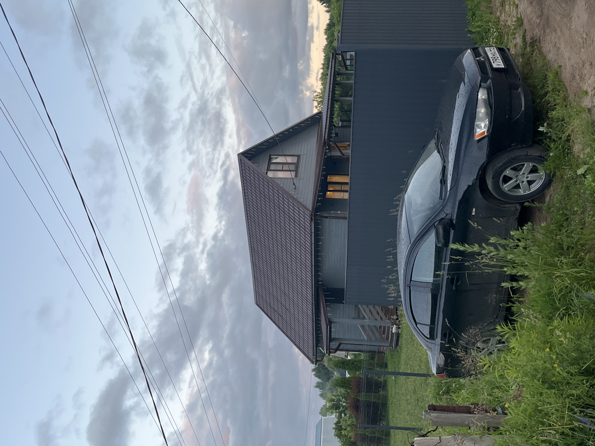 A black sedan is parked on a dirt and grass area in front of a grey wooden house with a dark brown roof in the village of Klyubushnevo, Russia. The house has windows and lights on, and there is a covered porch with string lights. A dark grey metal fence runs along the front of the property. The sky is cloudy and overcast, with power lines visible overhead. The car has a license plate that reads "Y798MA 44". The area is surrounded by greenery and trees.